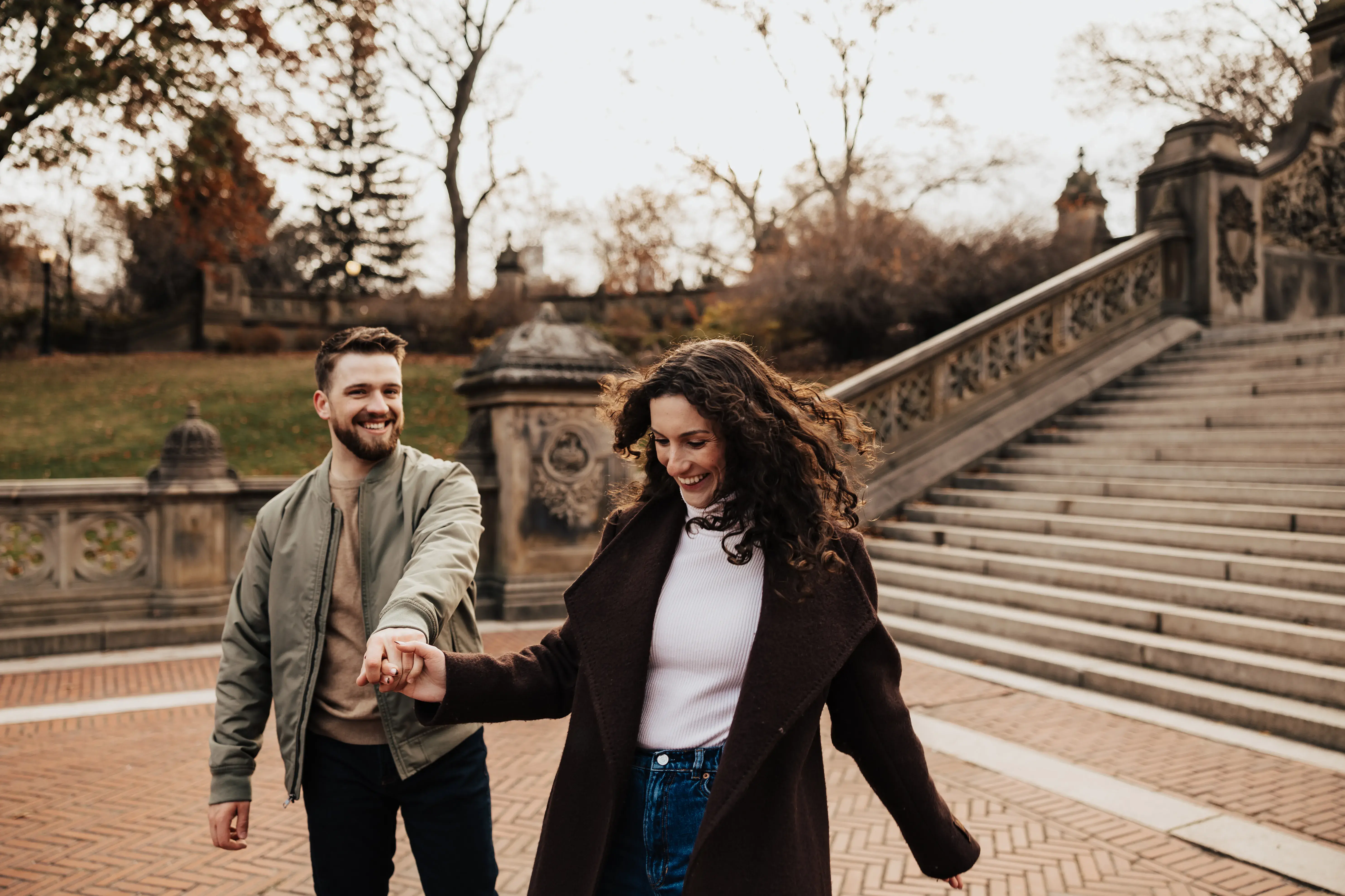 Couple holding hands and smiling as they walk beside the stone steps and ornate terrace in Central Park during a relaxed engagement session.