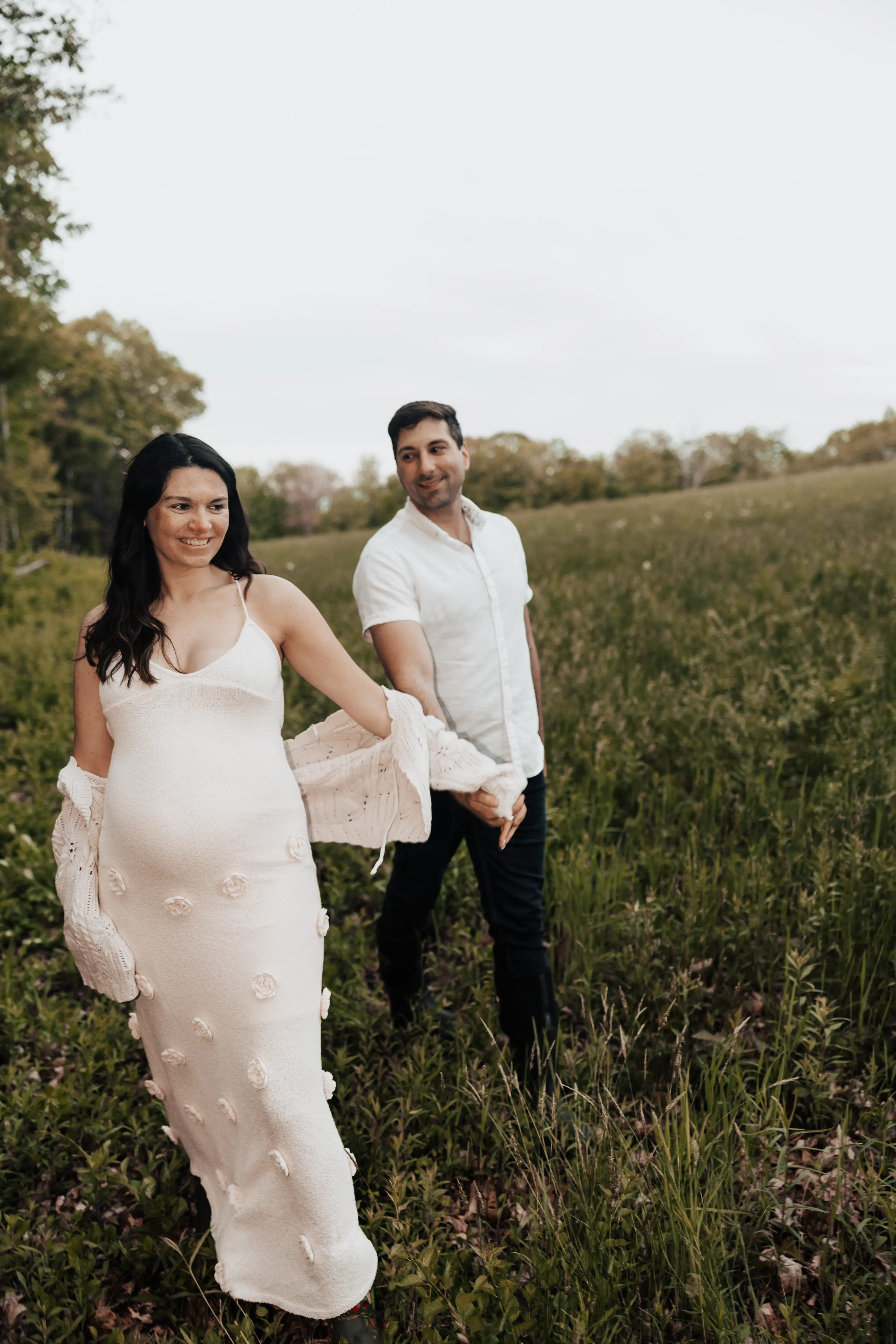 Expectant couple standing together in a quiet open field, holding hands and smiling as they share a peaceful maternity moment surrounded by trees and tall grass.