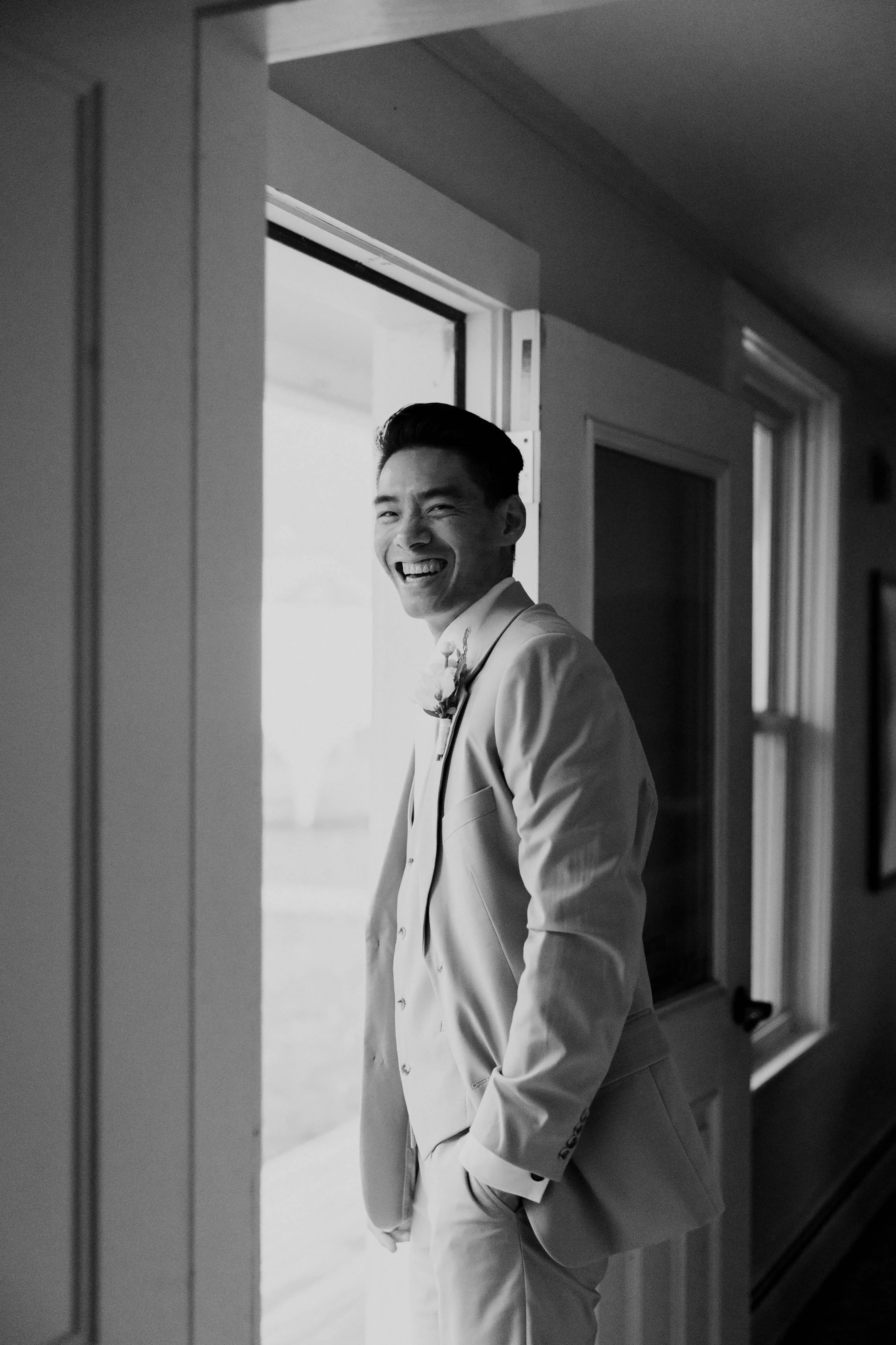 Groom smiling in a light-colored suit while standing in a doorway, captured in a candid black-and-white wedding portrait.