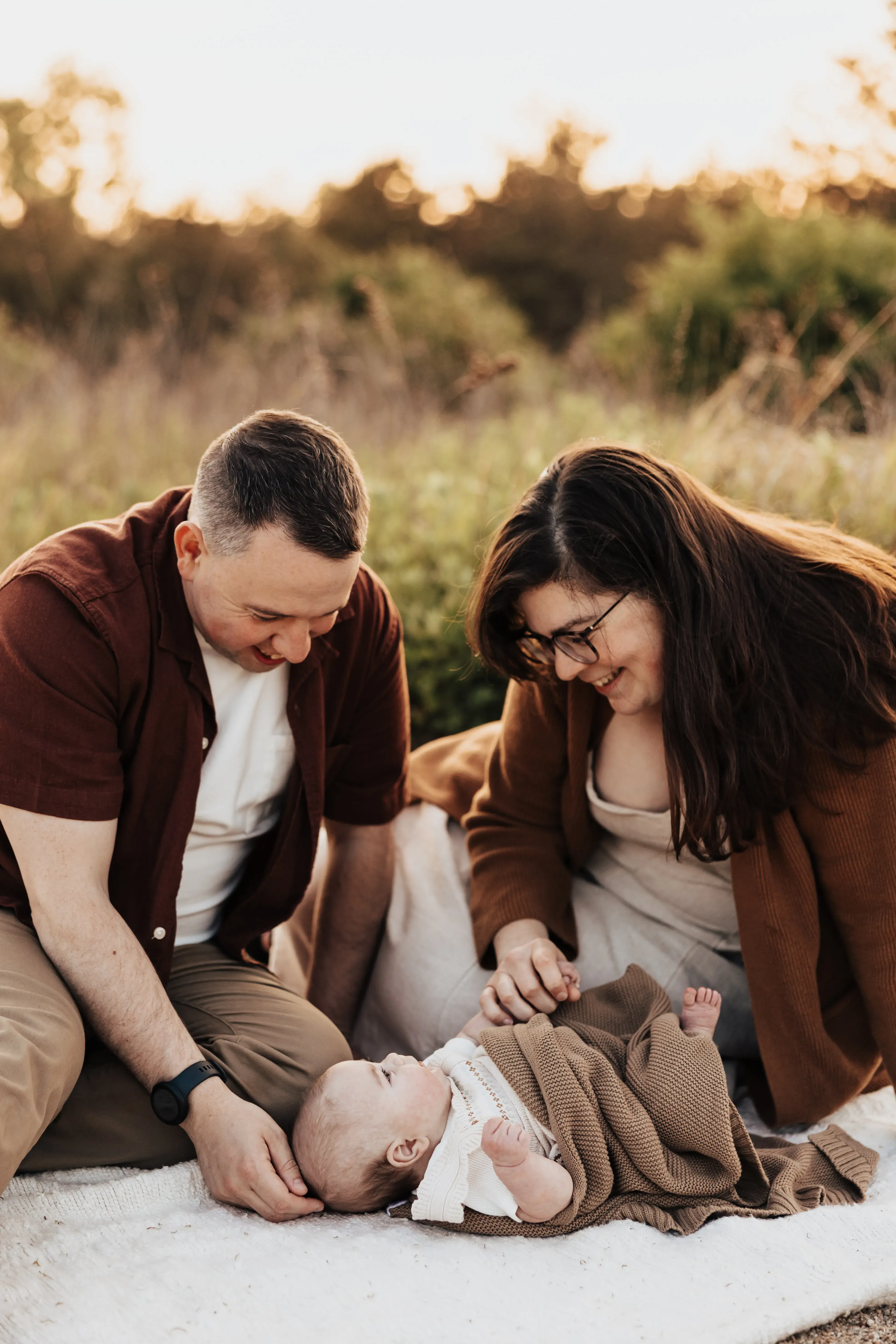 Parents smiling down at their newborn during a warm outdoor family session at sunset, with the baby wrapped in a soft brown blanket on a picnic blanket.