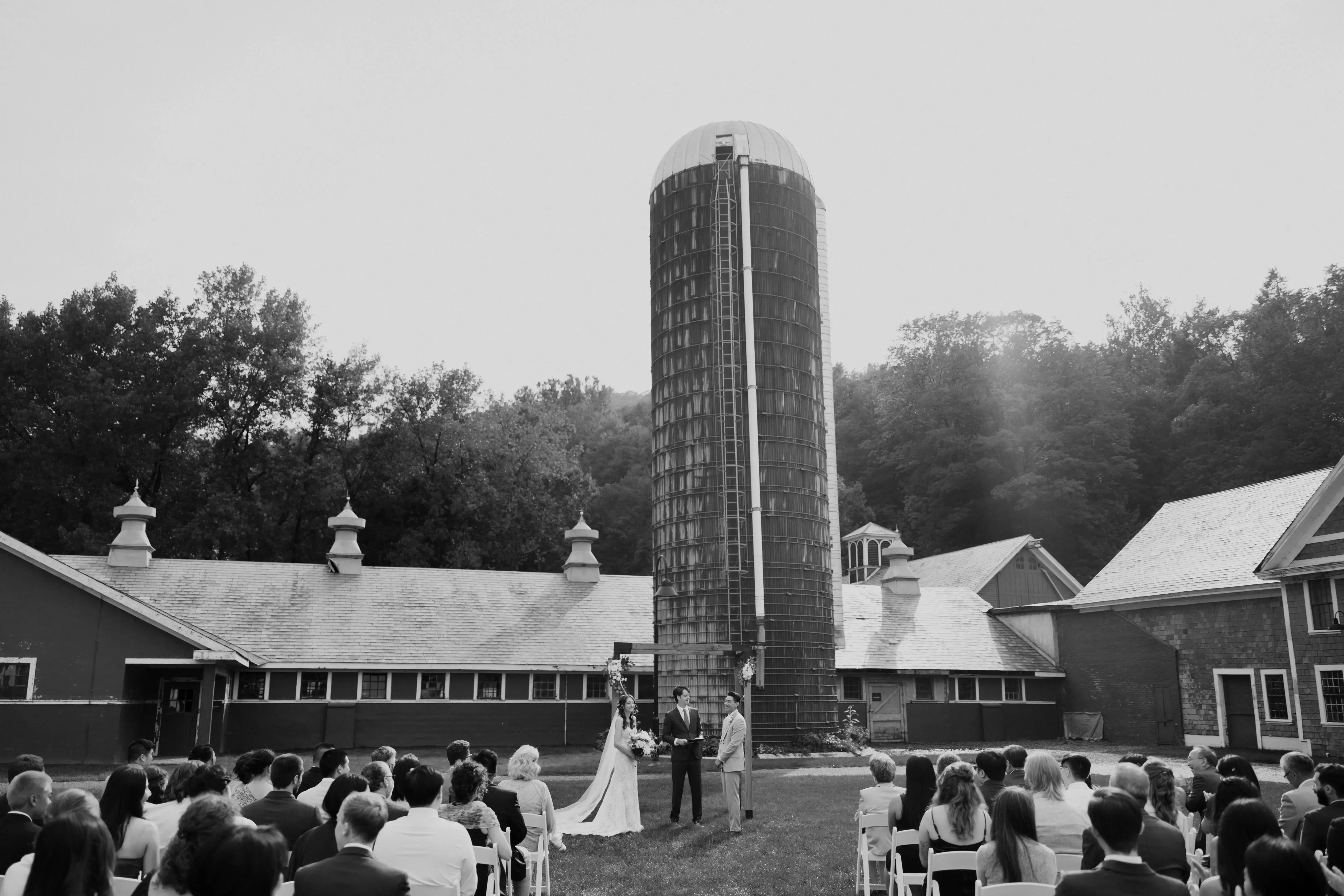 Bride and groom exchanging vows during an outdoor ceremony at a rustic farm venue, surrounded by seated guests with a tall silo rising behind them.