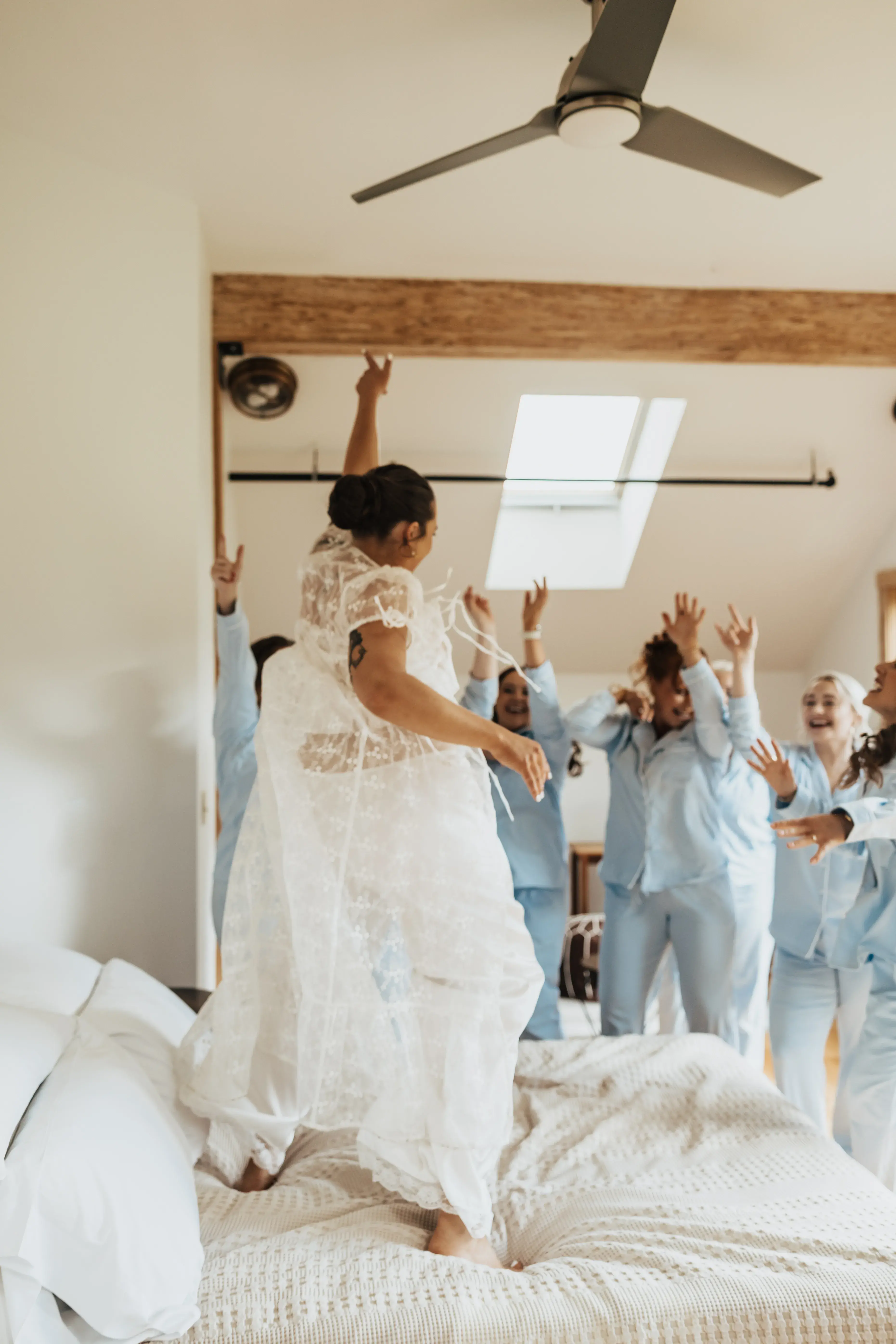 Bride laughing and dancing on the bed while her bridesmaids cheer around her in matching blue pajamas during a lively wedding morning getting-ready moment.