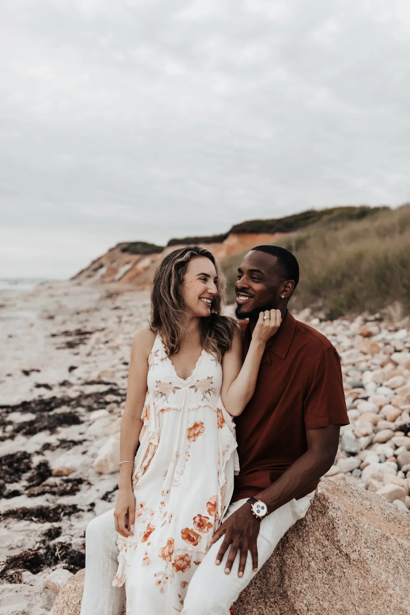 Couple sitting together on a rocky beach, smiling at each other during a relaxed coastal portrait session with dunes and ocean shoreline in the background.
