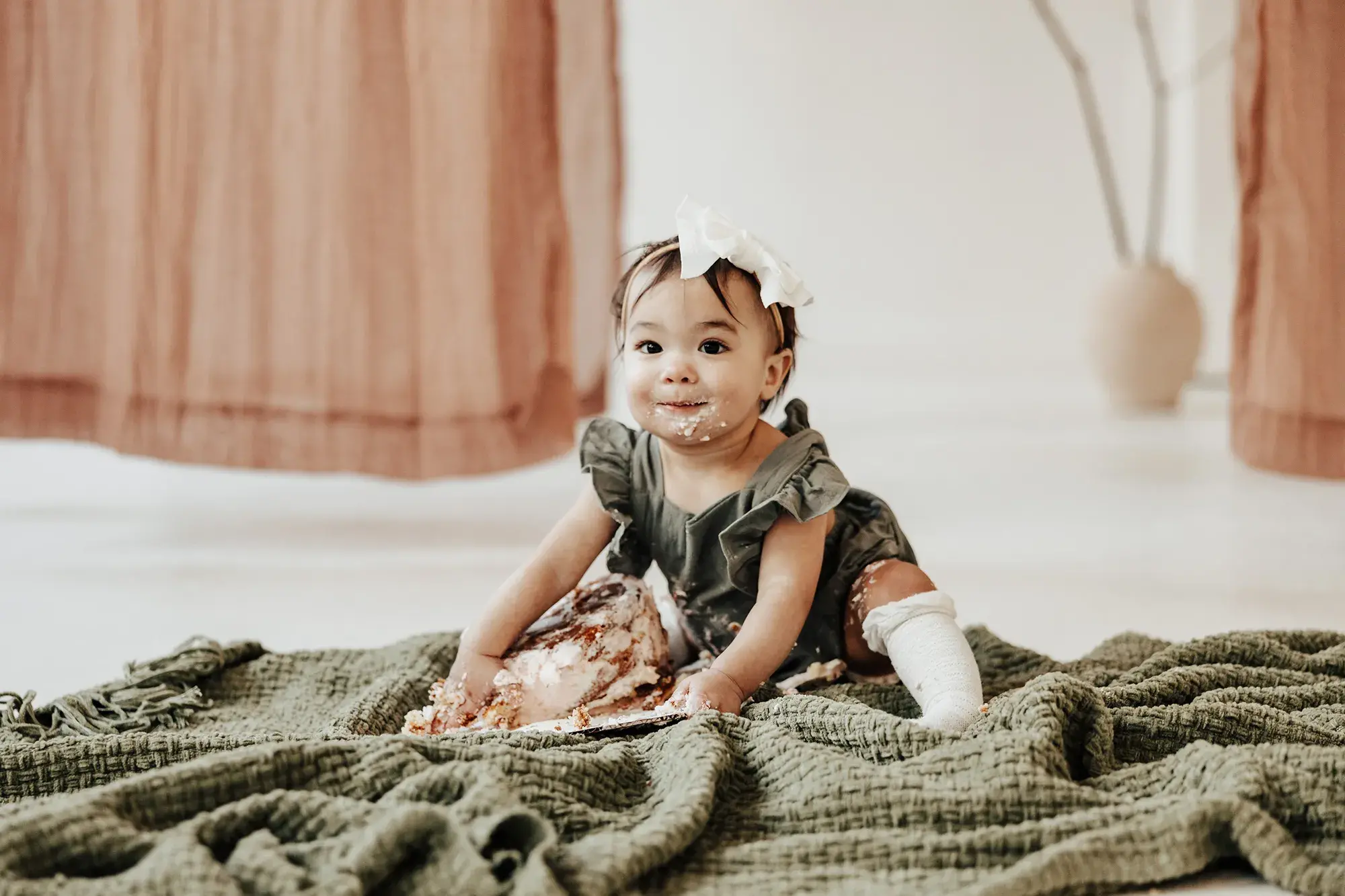 Baby smiling with cake and frosting on her face during a playful first birthday cake smash session, sitting on a soft blanket in a bright studio setting.