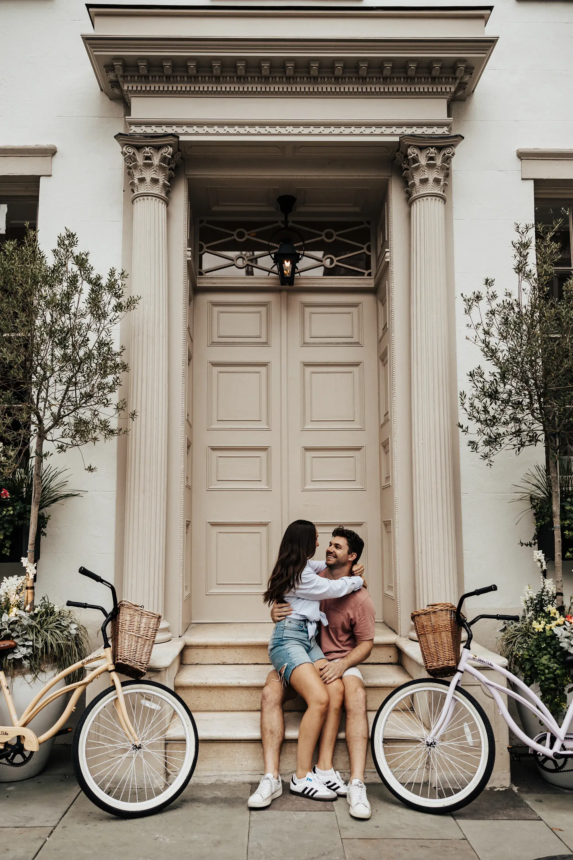 Couple sitting close together on the front steps of an elegant home, smiling and embracing between two vintage bicycles with wicker baskets.