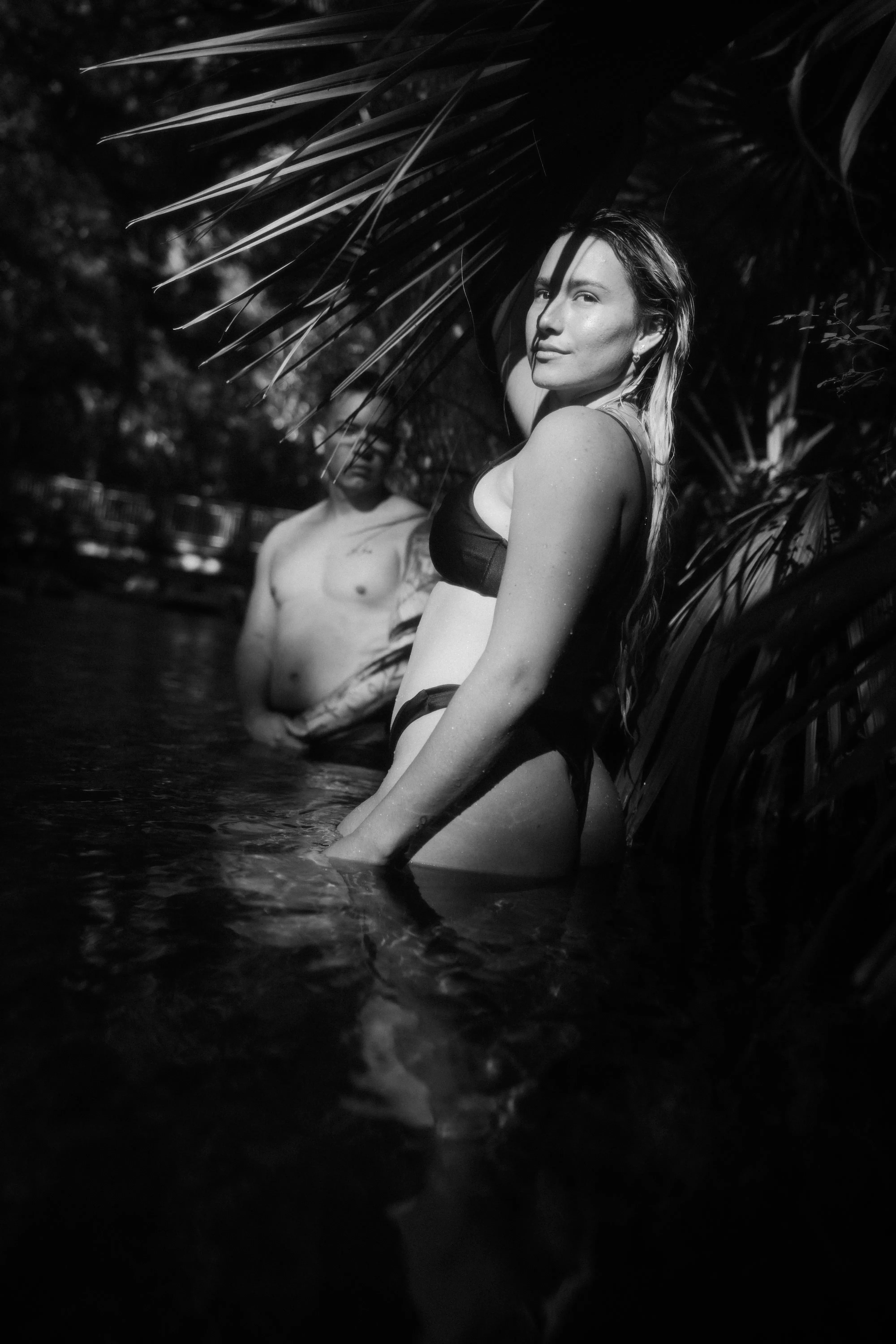 Couple standing waist-deep in a shaded tropical spring, framed by palm leaves in a dramatic black-and-white portrait.