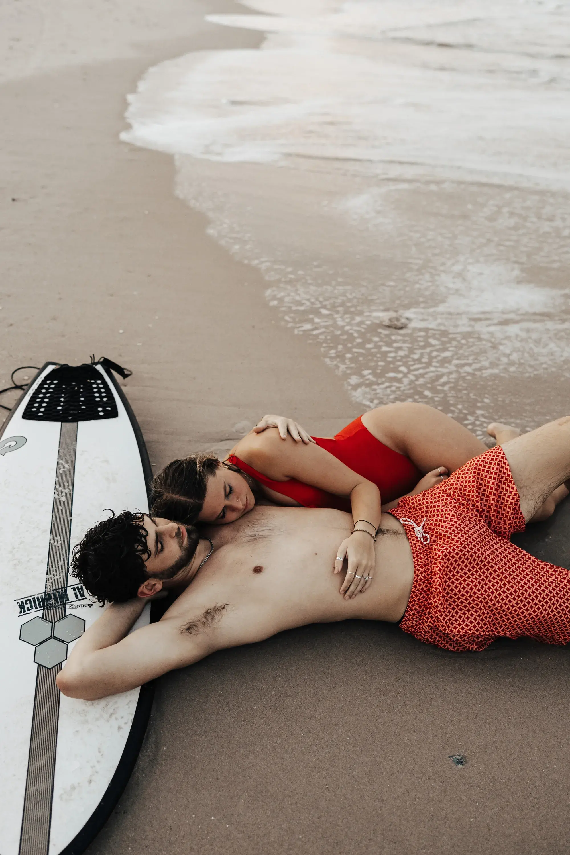 Couple relaxing together on the beach beside a surfboard, sharing a quiet moment near the shoreline as gentle waves wash onto the sand.