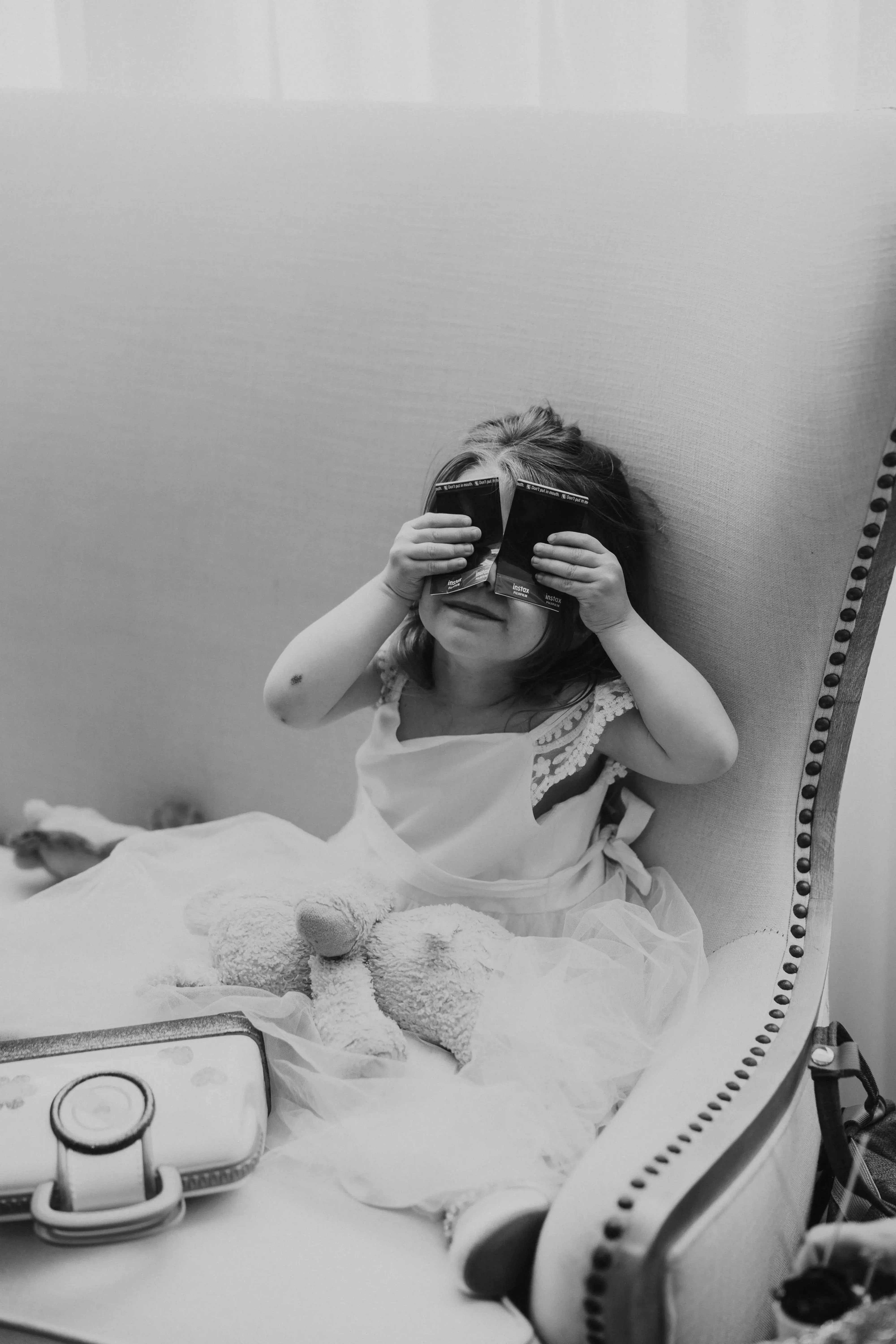 Young flower girl sitting in a cushioned chair and playfully covering her eyes with instant photo prints during a candid black-and-white wedding moment.