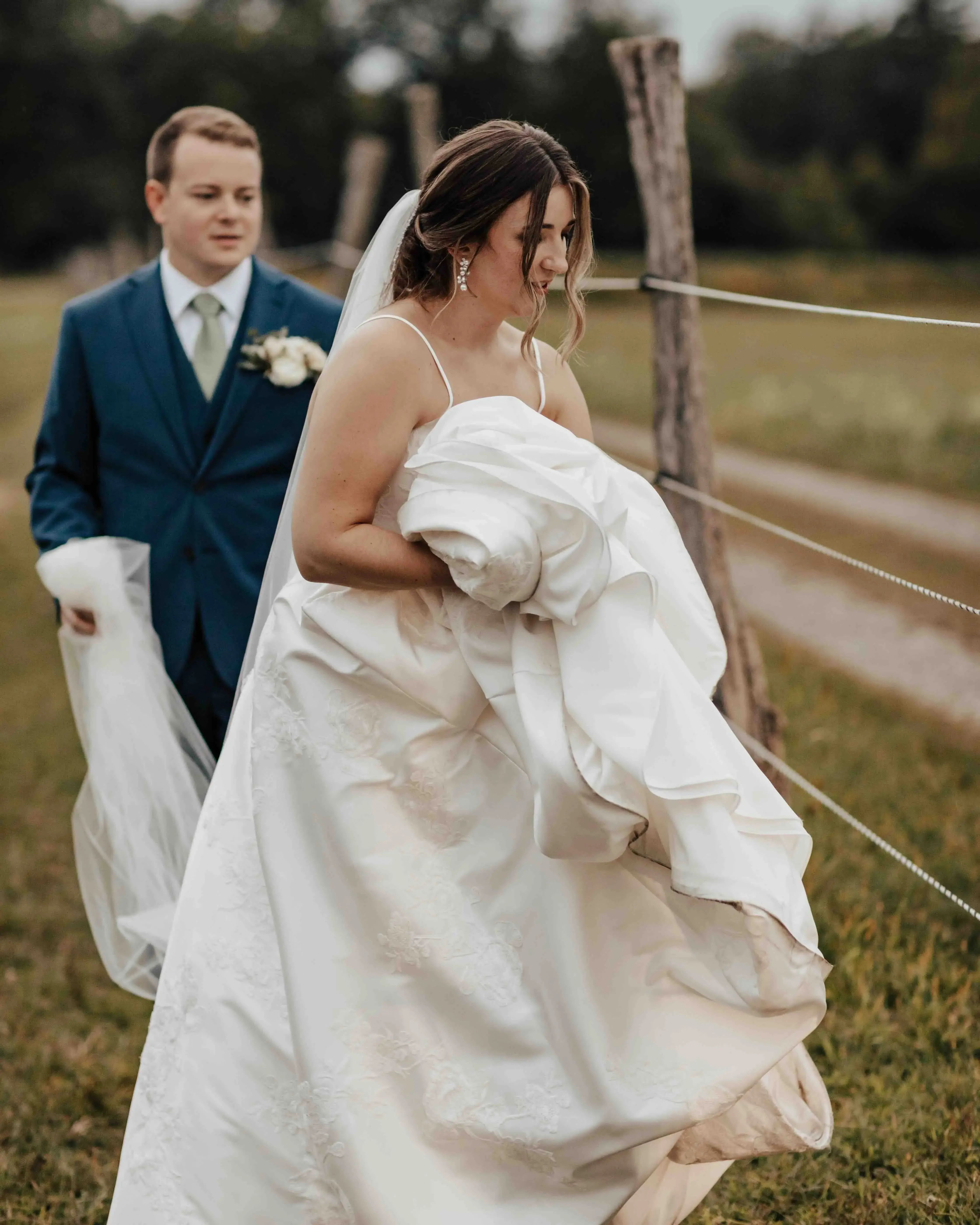 Bride gathering the train of her wedding dress as she walks through a grassy field, with the groom following behind and carrying part of her veil.