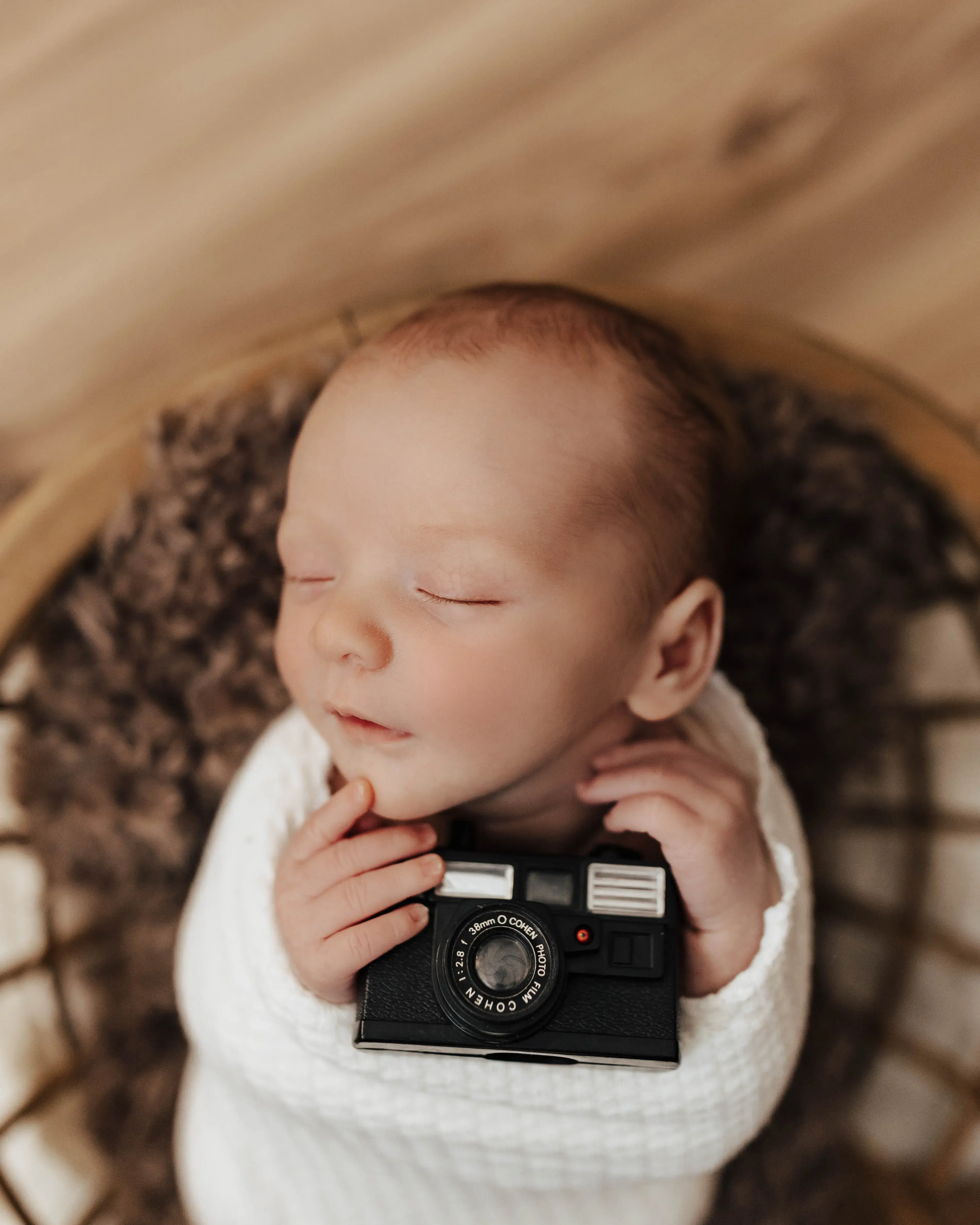 Newborn baby sleeping peacefully while wrapped in a soft white blanket and gently holding a small vintage camera during a cozy studio portrait.