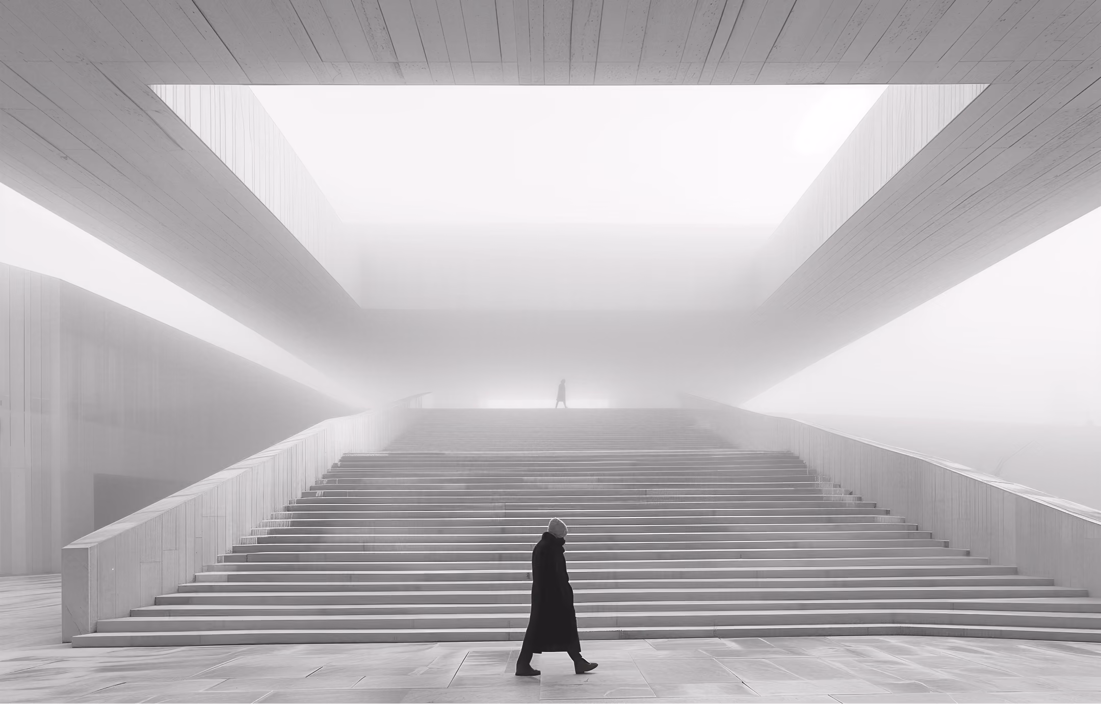 Person in dark coat walking in front of wide staircase inside a foggy modern architectural space.
