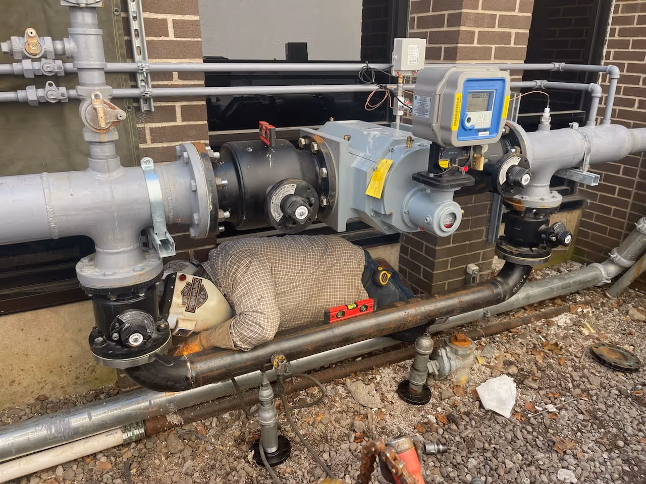 Worker wearing plaid shirt and welding helmet welding a pipe under industrial piping with gauges.