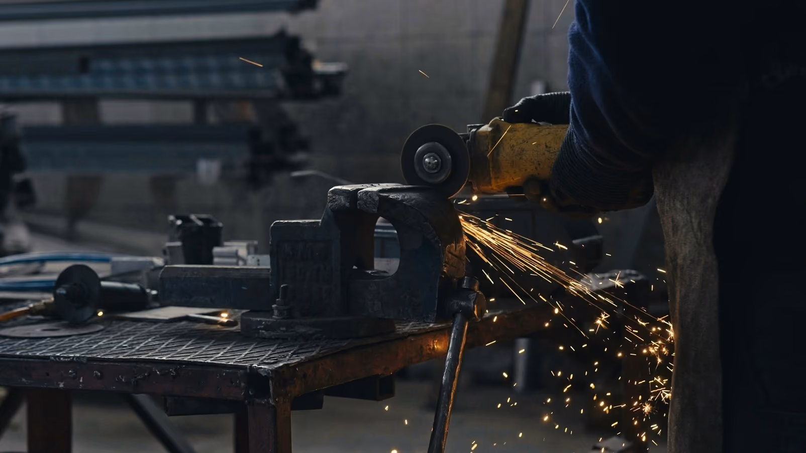 Worker using a power grinder on a metal piece clamped in a vise, with sparks flying in a workshop.