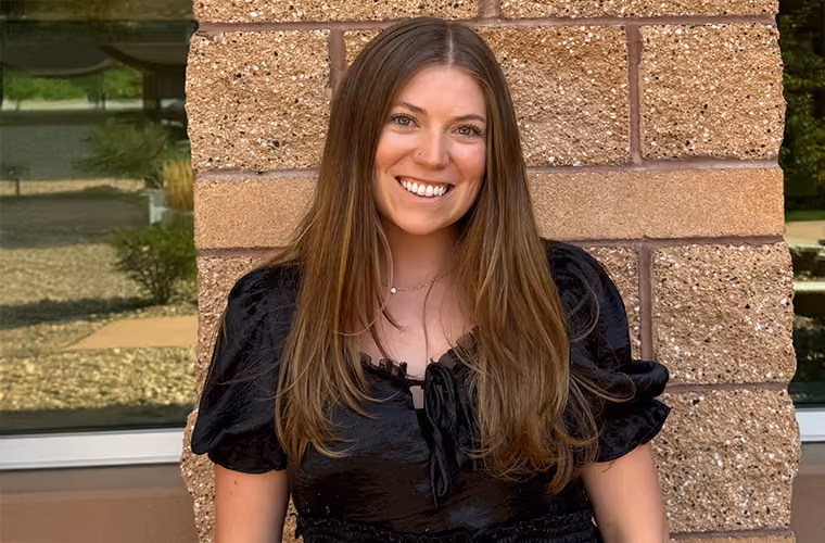 Headshot of a smiling woman with long brown hair in front of a brick wall.