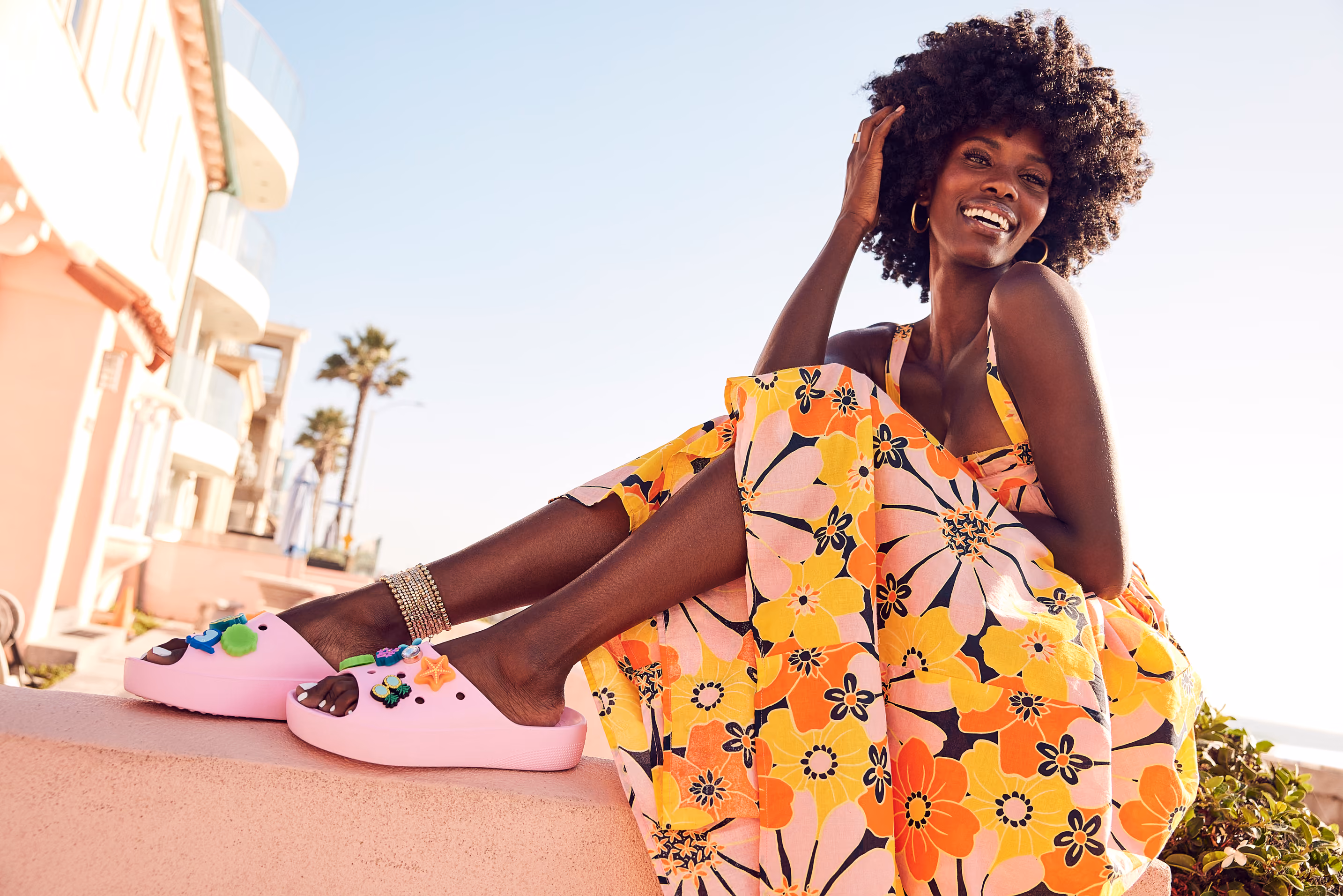 Smiling woman in a floral dress seated on a beachfront wall beside pink Crocs clogs.