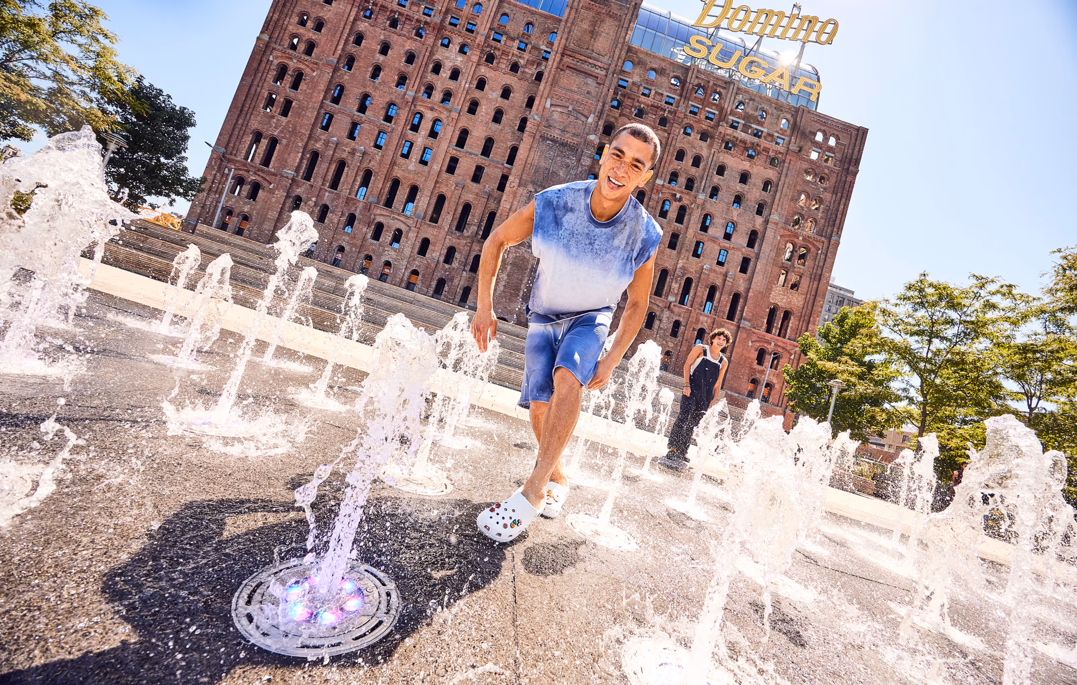 Young man playing in a city splash fountain wearing white Crocs clogs.