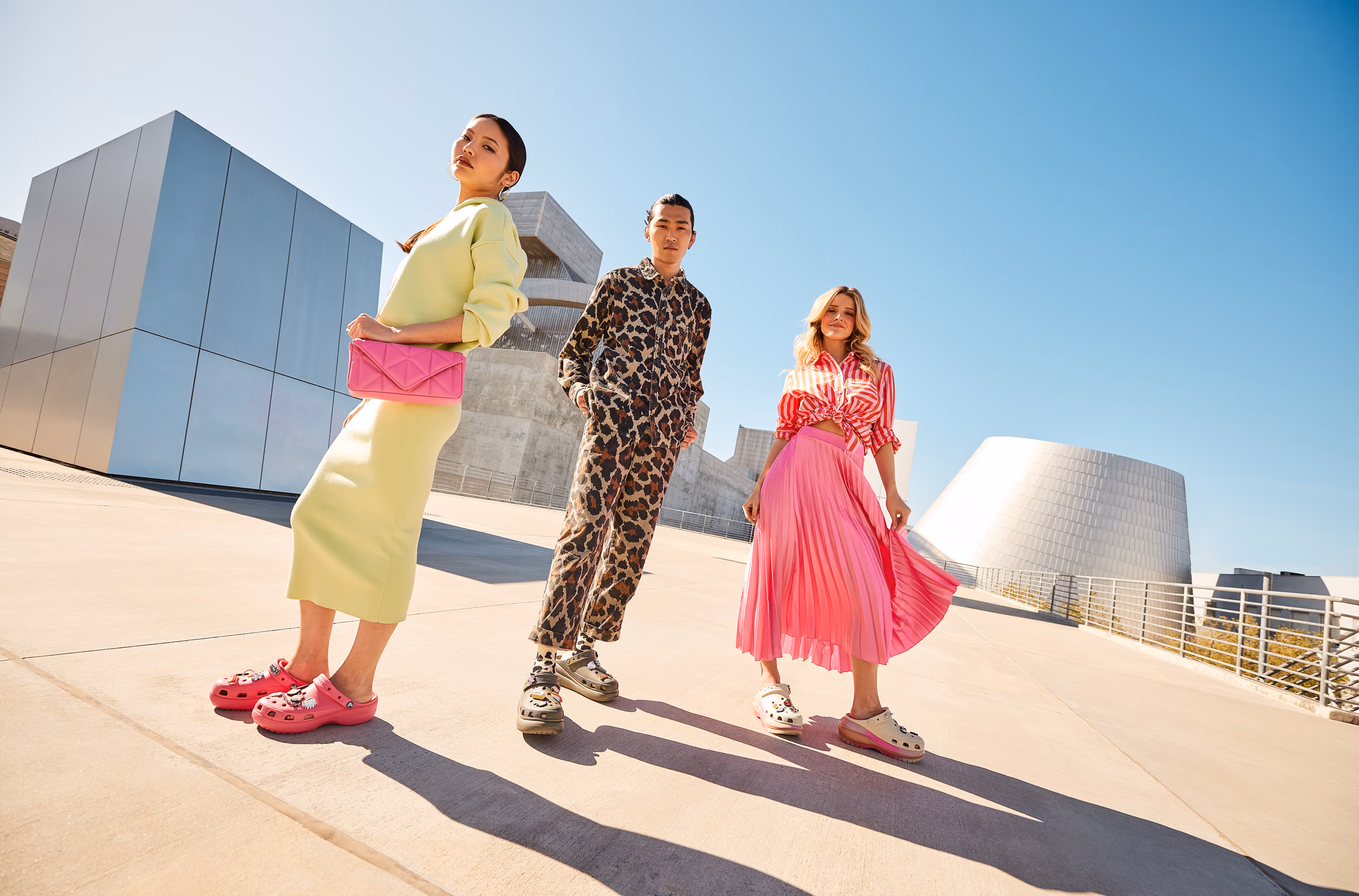 Three friends in colorful outfits walking by a modern building in Crocs platform clogs.