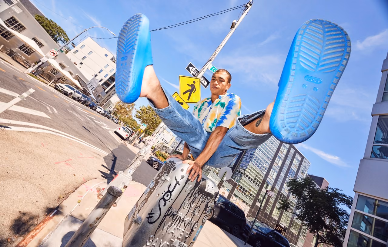 Man leaping on a skateboard showing the blue soles of his Crocs clogs in the city.
