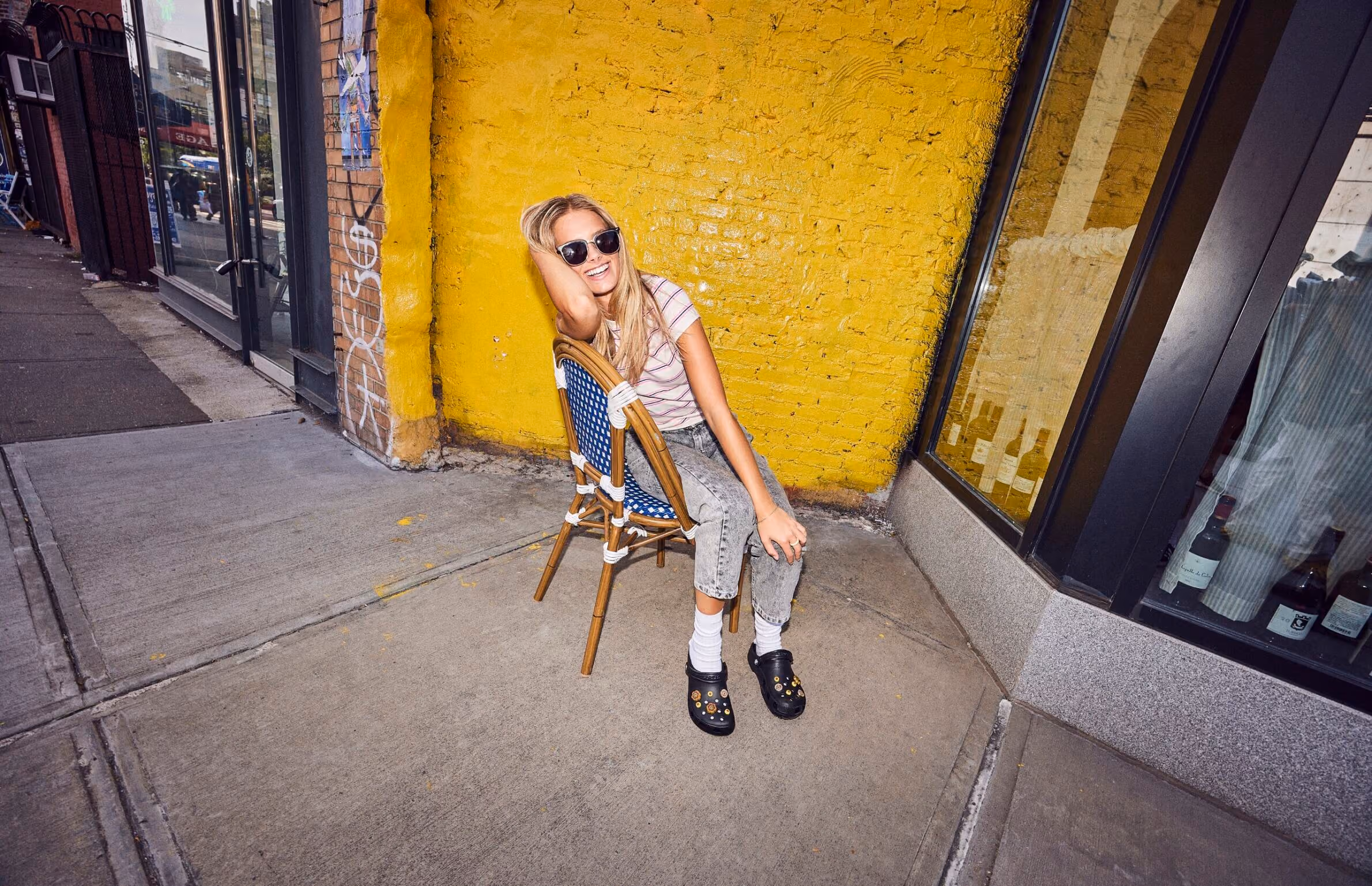 Woman sitting on a bistro chair against a yellow wall wearing black Crocs clogs.