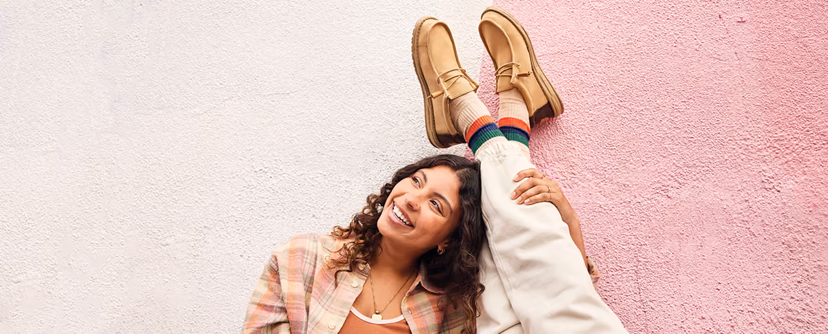 Smiling woman lying against a pink wall with her feet up, wearing tan HEYDUDE boots.