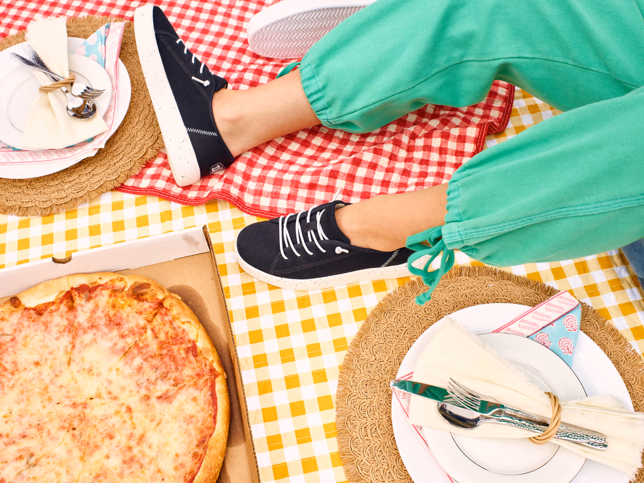 Overhead view of navy HEYDUDE sneakers beside a pizza on a checkered picnic blanket.