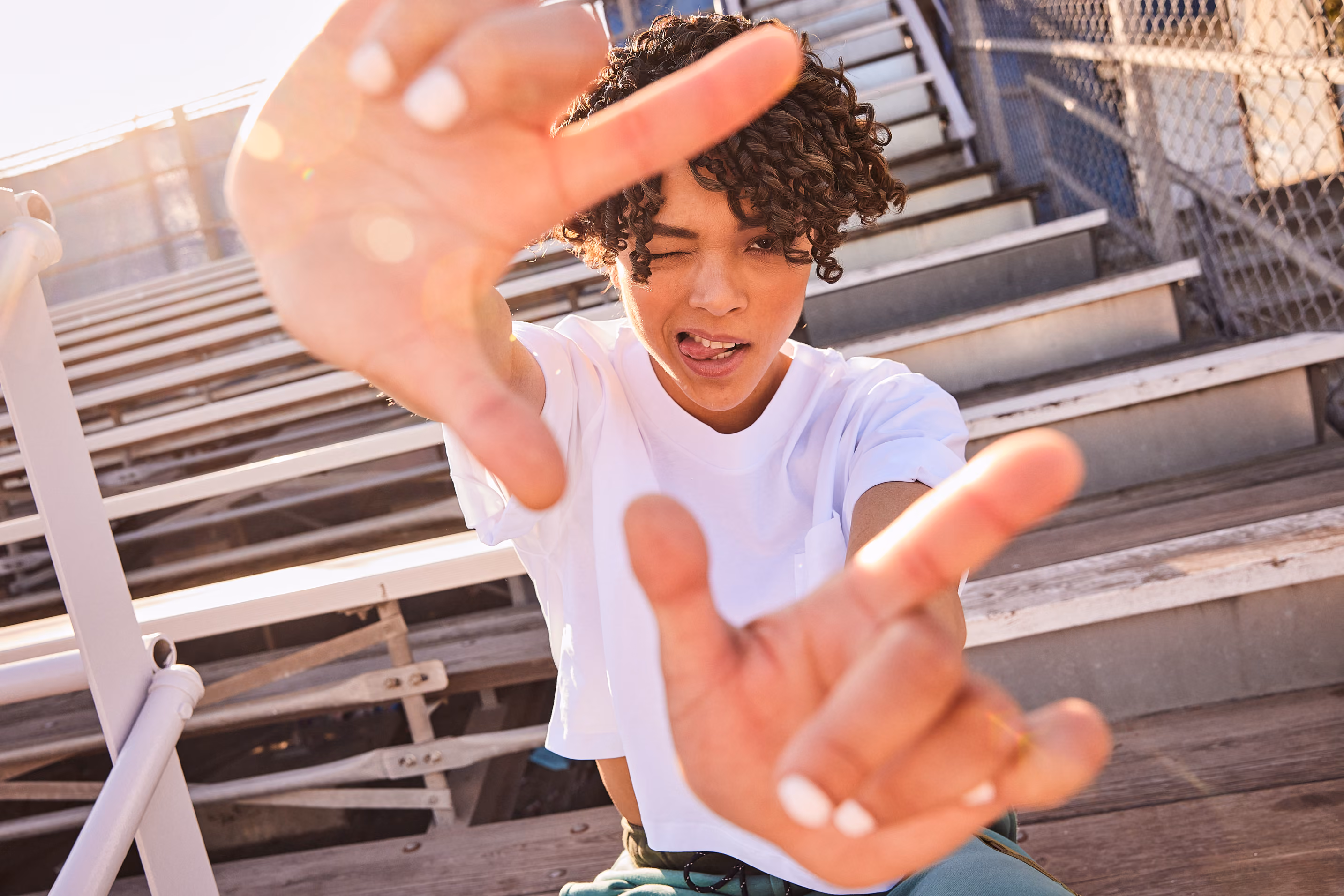 Young person on stadium bleachers reaching toward the camera in a white tee.