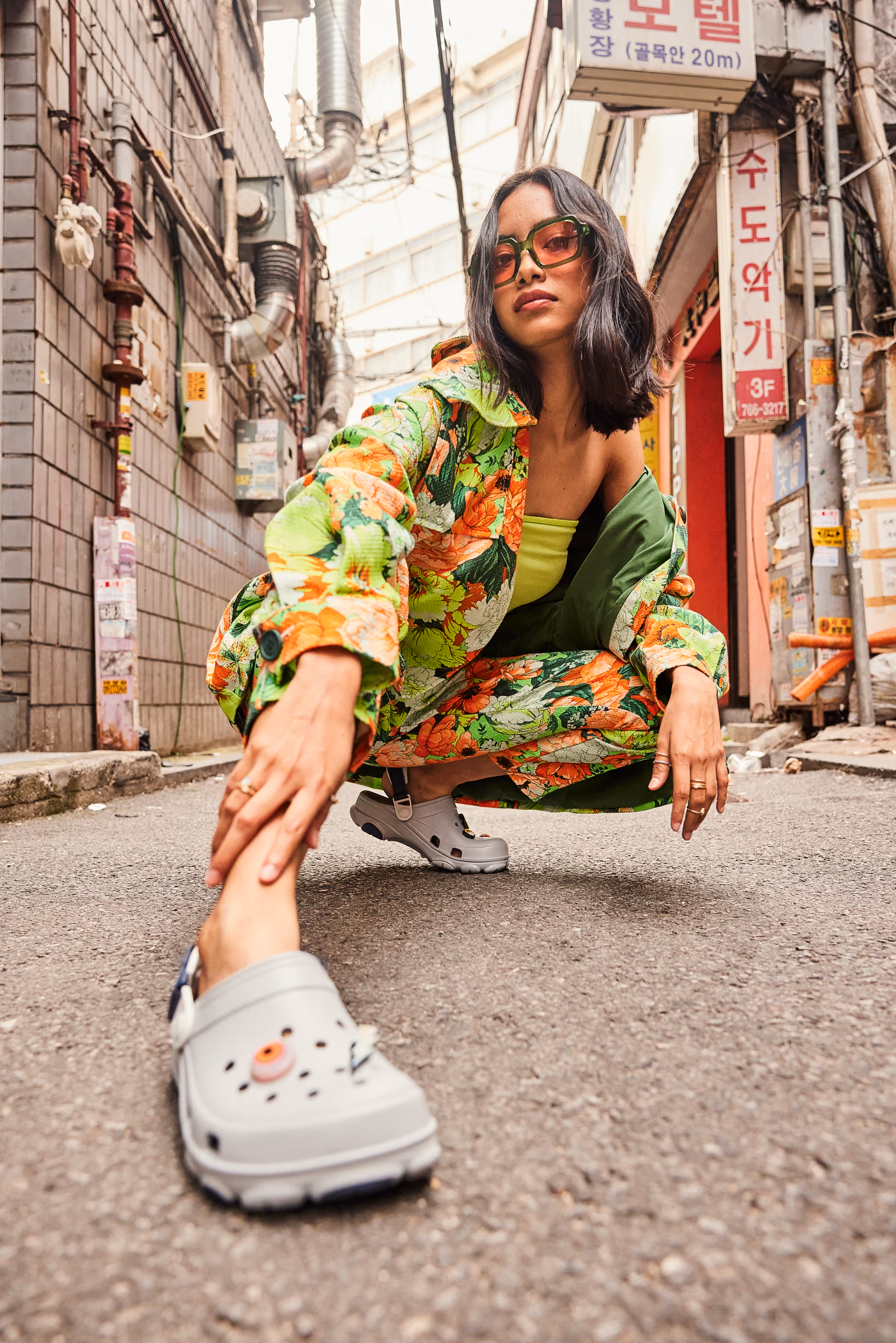 Woman crouching in a narrow city alley wearing a floral set and gray Crocs clogs.