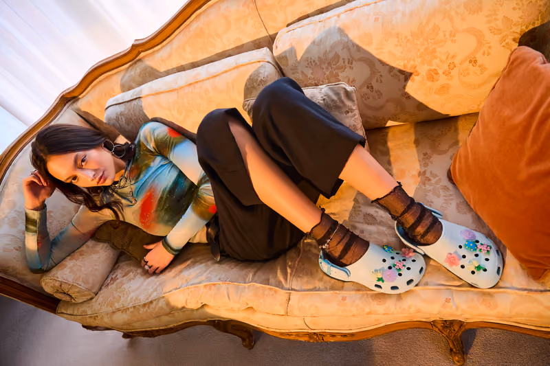 Woman reclining on a vintage sofa wearing white Crocs clogs with colorful Jibbitz charms.
