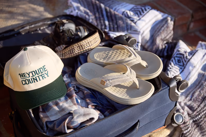 HEYDUDE shoes, sandals, and a branded cap arranged on a blanket.