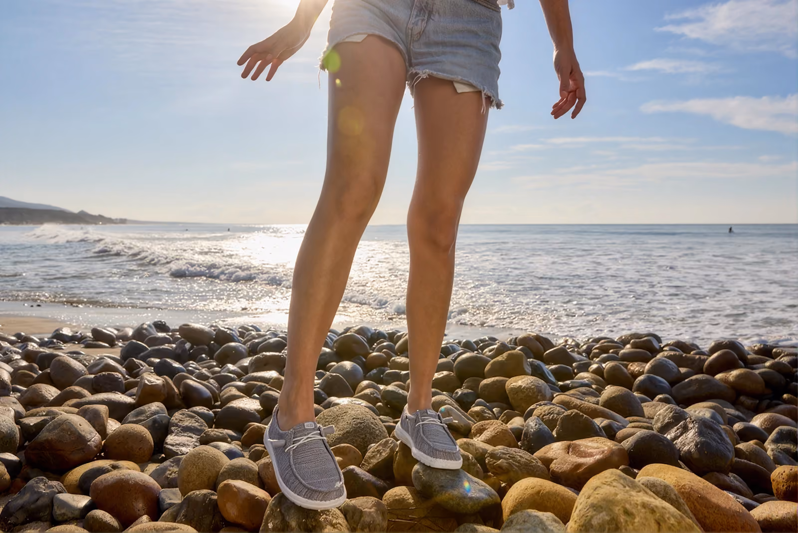Person standing on a rocky beach wearing gray HEYDUDE shoes.