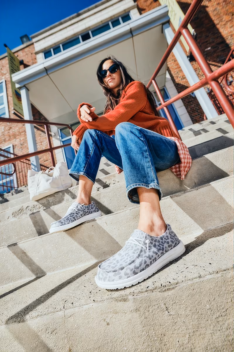 Woman sitting on outdoor steps wearing patterned HEYDUDE slip-on shoes.