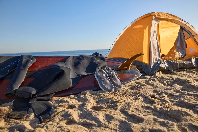 Camping gear and shoes beside an orange tent on a sandy beach.
