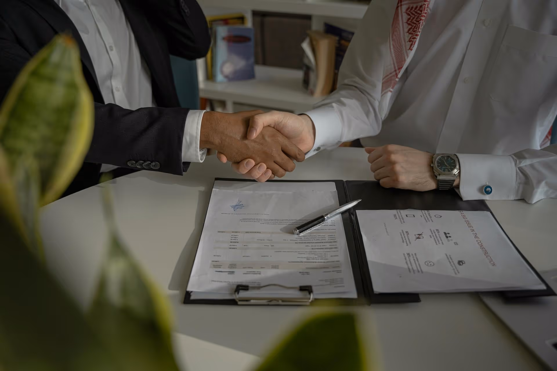 Two businesspeople shaking hands over paperwork on a desk, with documents and a pen visible in a professional office setting.