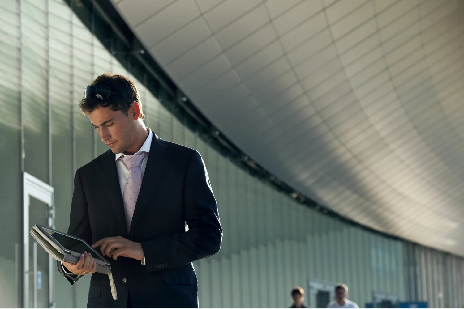 A man in a suit and tie is standing outside a modern building, holding a tablet. He has sunglasses perched on his head and appears to be focused on the screen of the tablet. The background features a curved roof and large windows, with a few people visible in the distance.