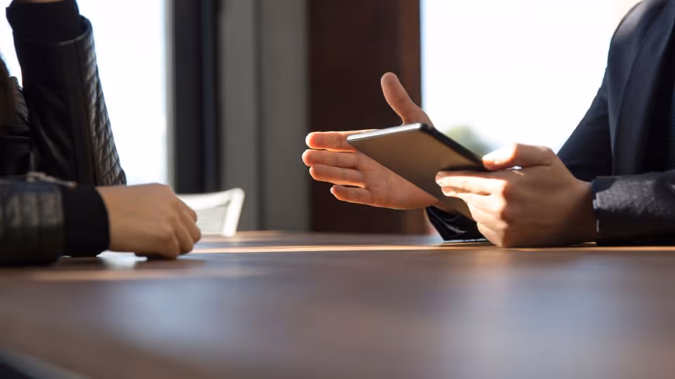 Two people sitting at a wooden table, one holding a tablet and gesturing with their hand.