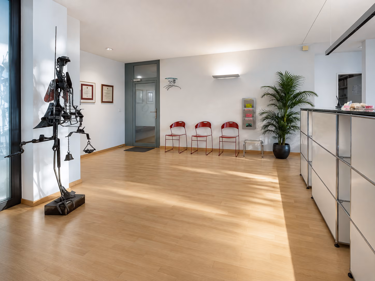 Heller Warteraum mit drei roten Stühlen, grünem Pflanzenkübel und moderner Skulptur aus Metall.