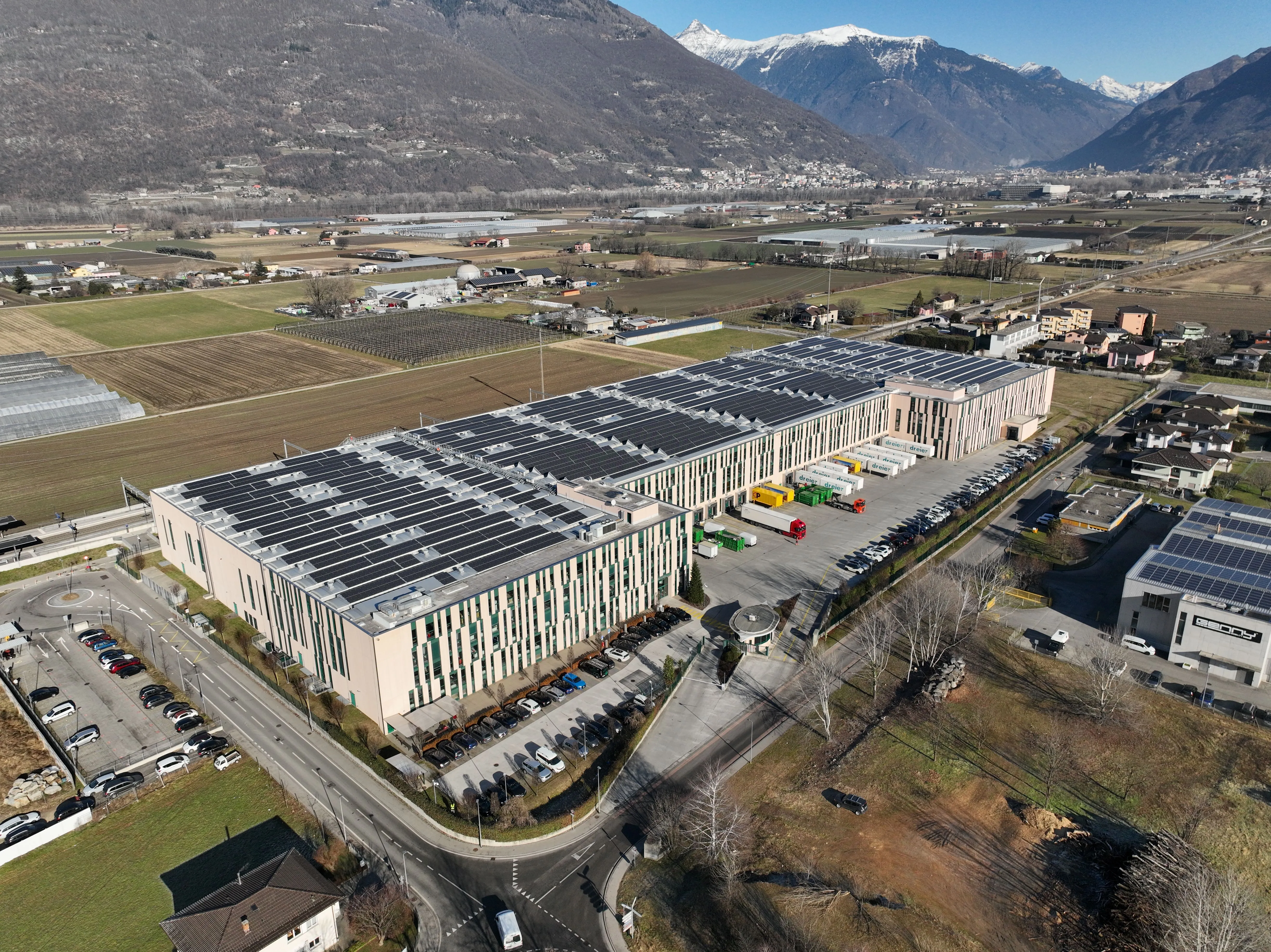 Aerial view of a large solar-powered industrial facility surrounded by farmland and snow-capped mountains.
