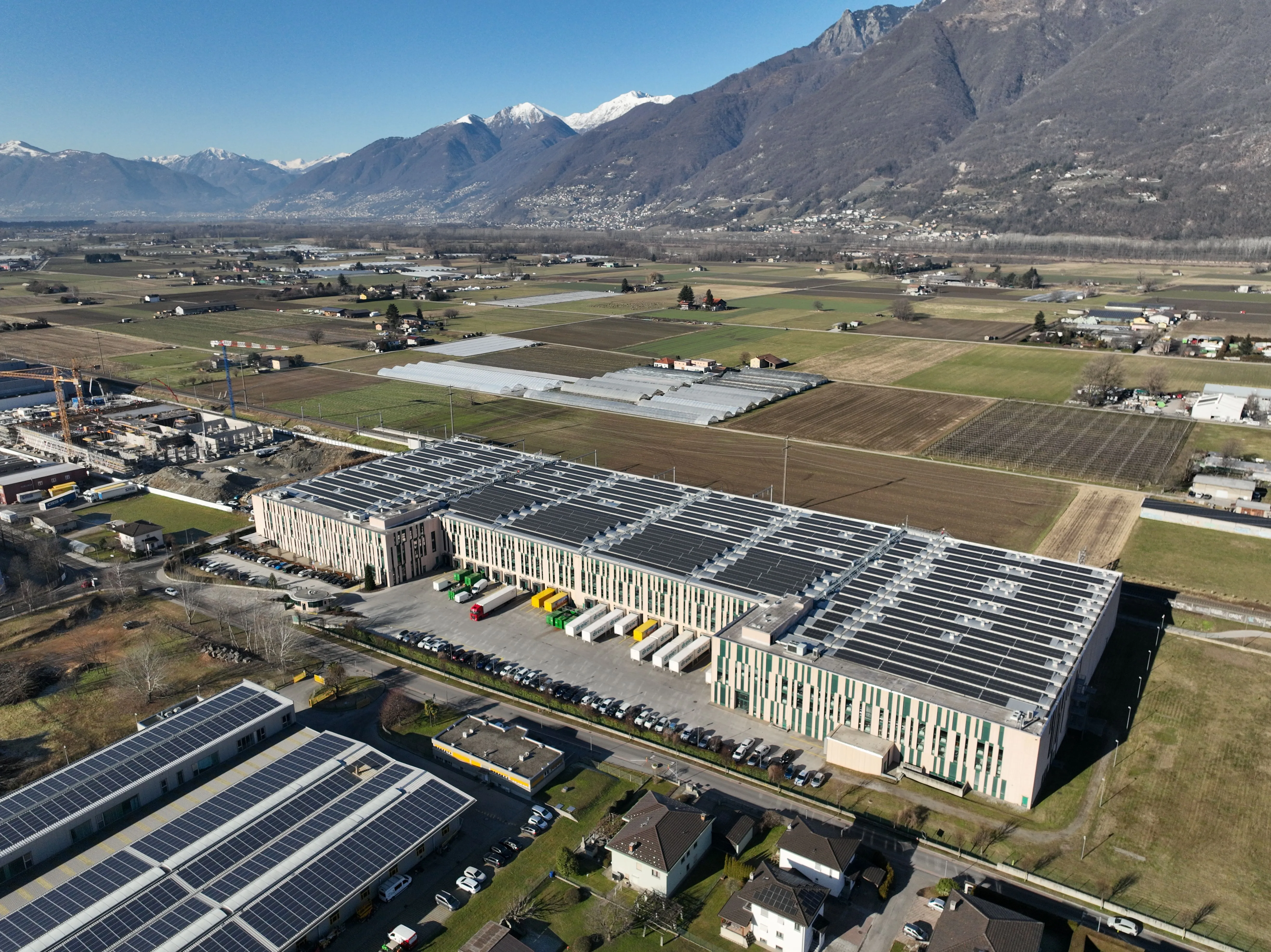 Aerial view of a large industrial facility with rooftop solar panels, surrounded by farmland and mountains.
