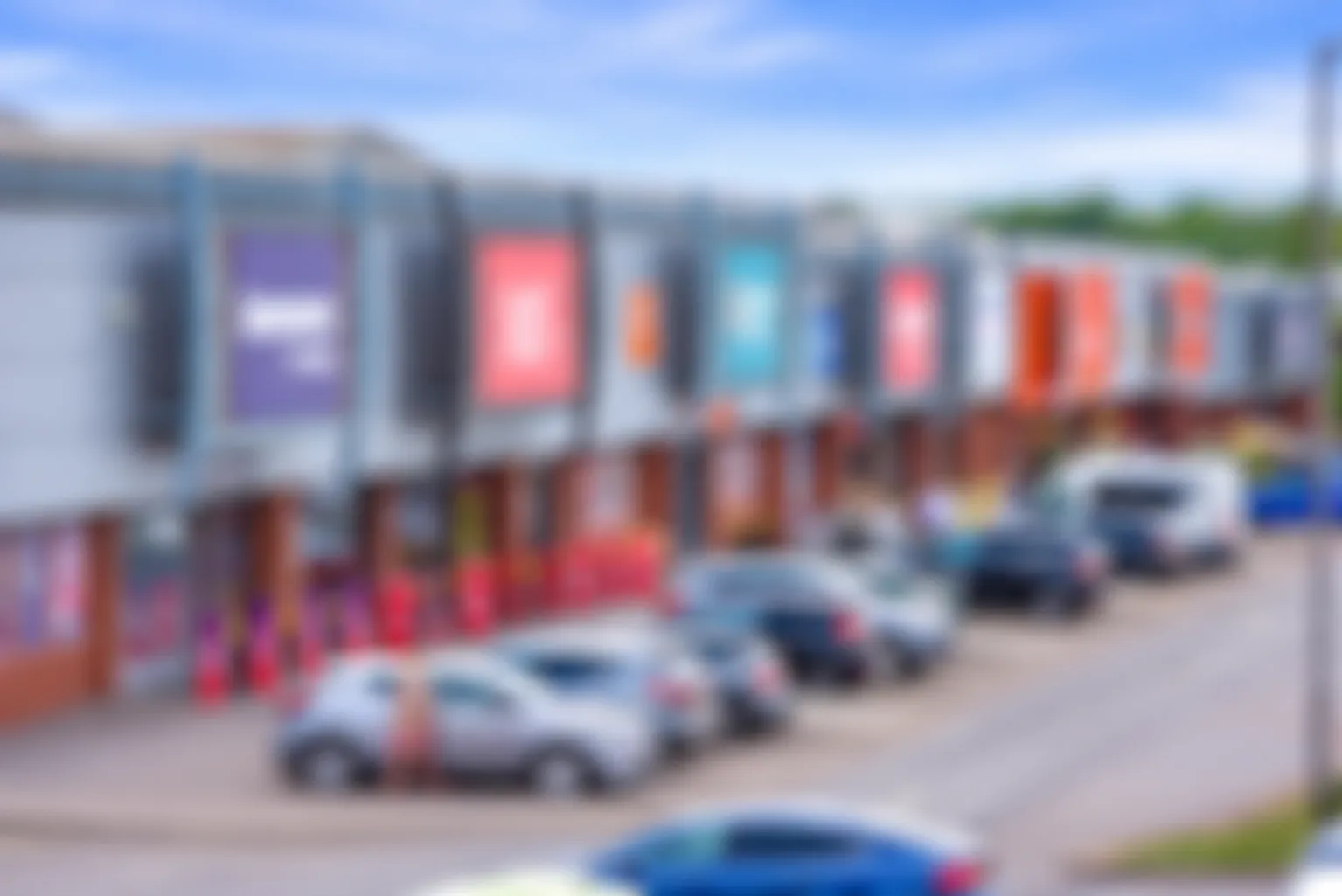 Blurred view of a retail shopping center with colorful storefronts and parked cars.
