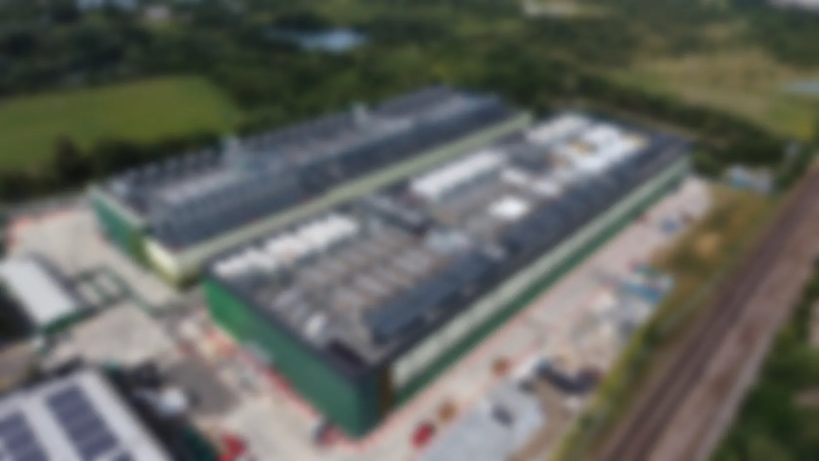 Blurred aerial view of a large industrial facility with green surroundings and adjacent railway tracks.