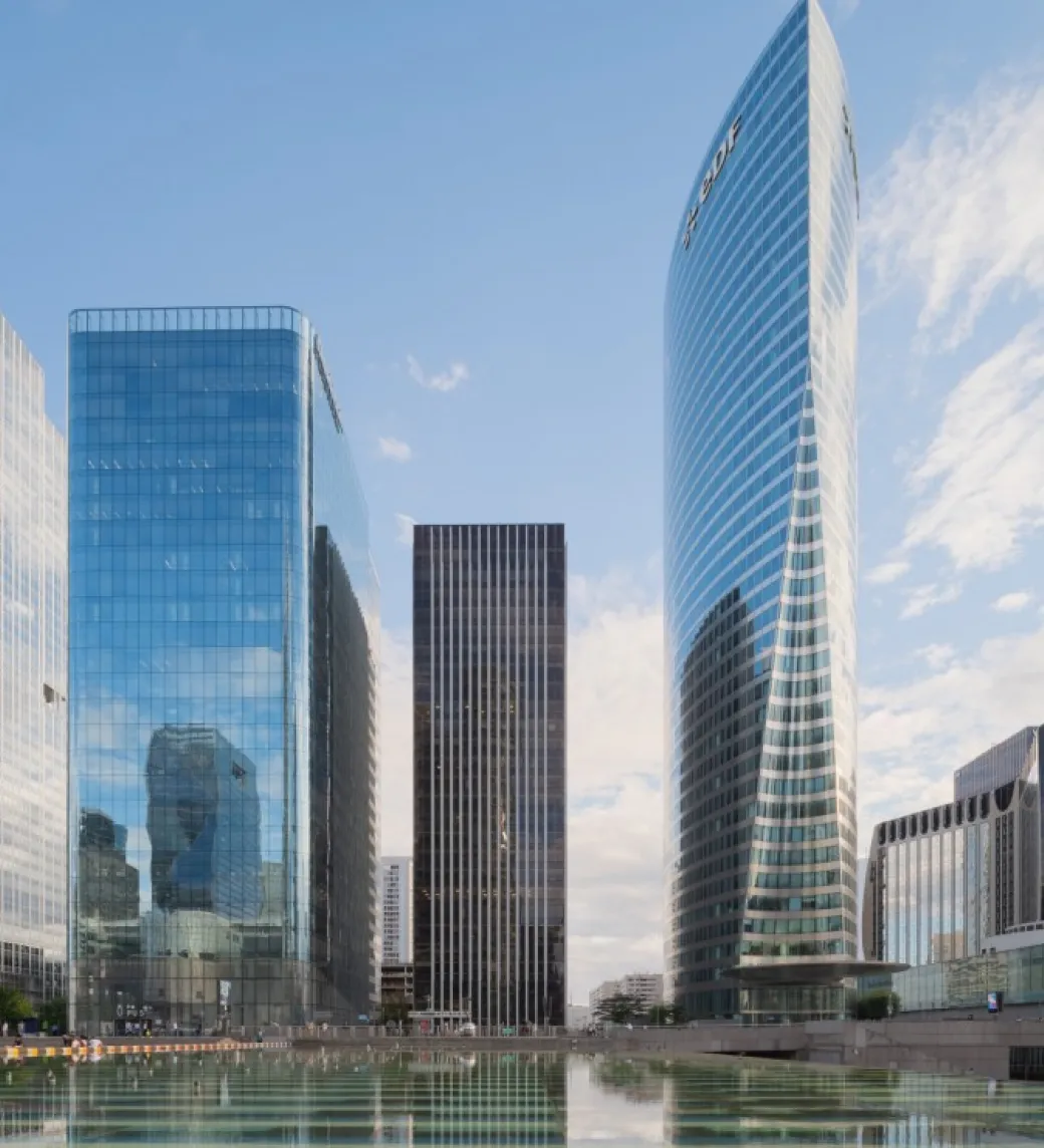 Modern glass skyscrapers reflecting blue sky and clouds above a calm water surface.