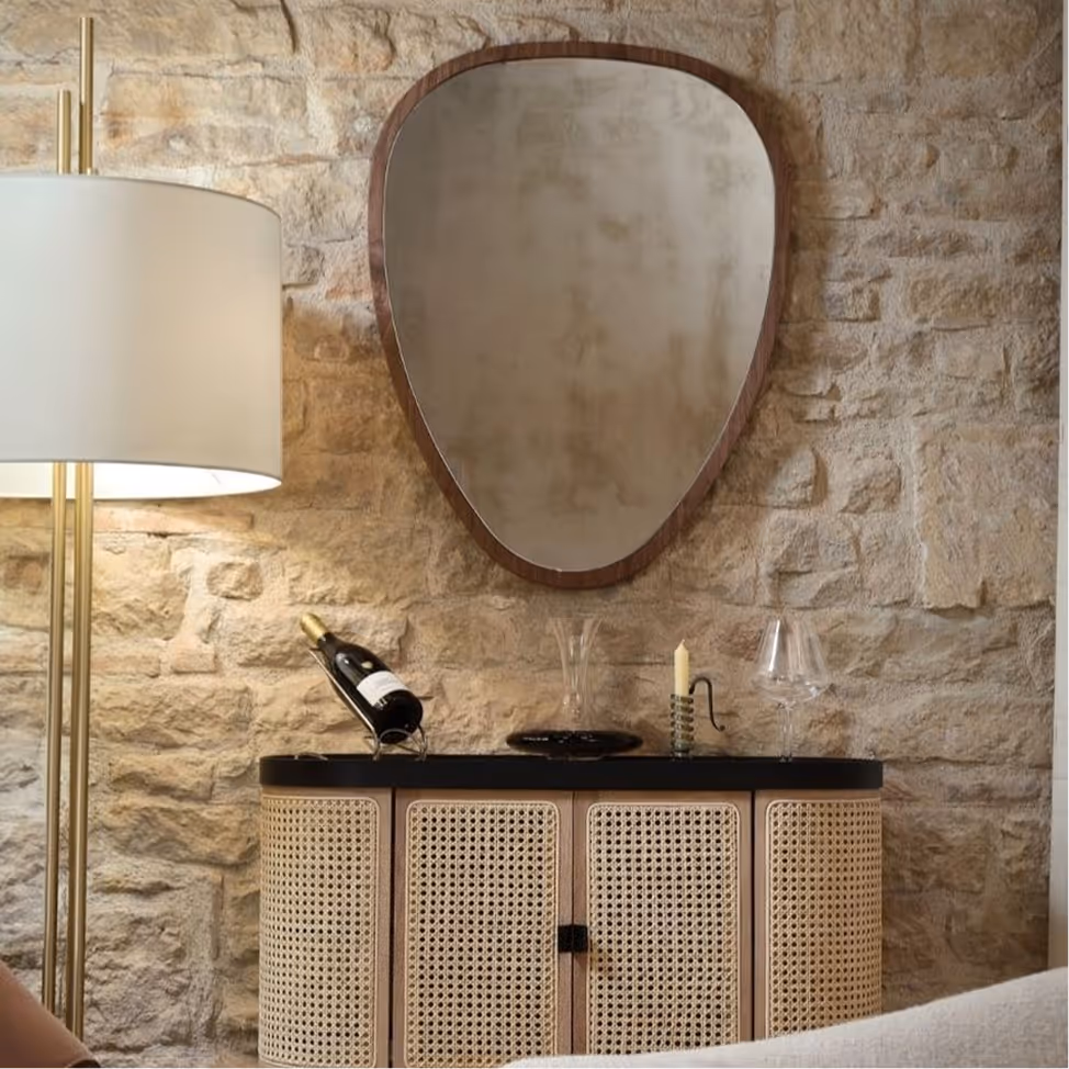 Stone wall with oval wooden-framed mirror above a cabinet holding a wine bottle, candle, and glass.