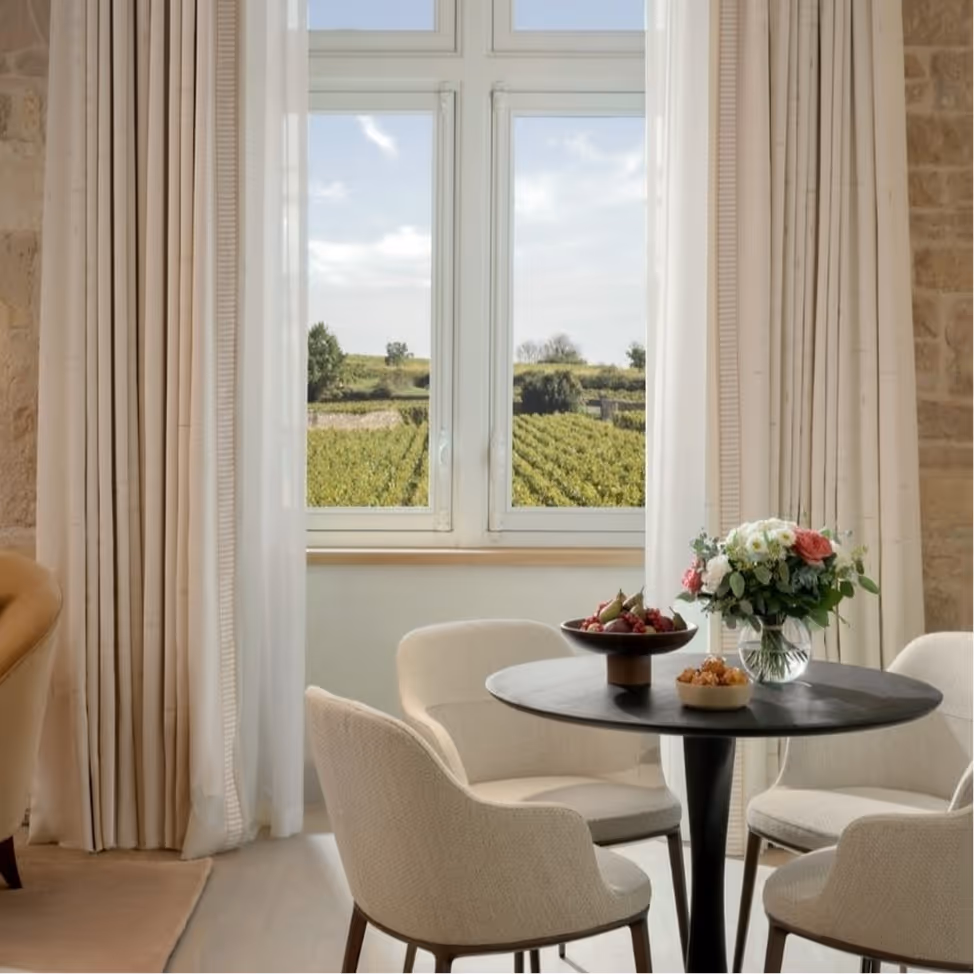 Cozy dining area with round table, four chairs, flowers, and window view of green farmland.