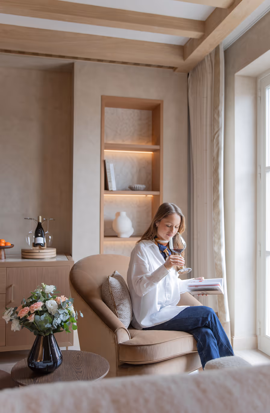 Woman sitting in beige armchair by window, holding wine glass and book in a softly lit modern living room.
