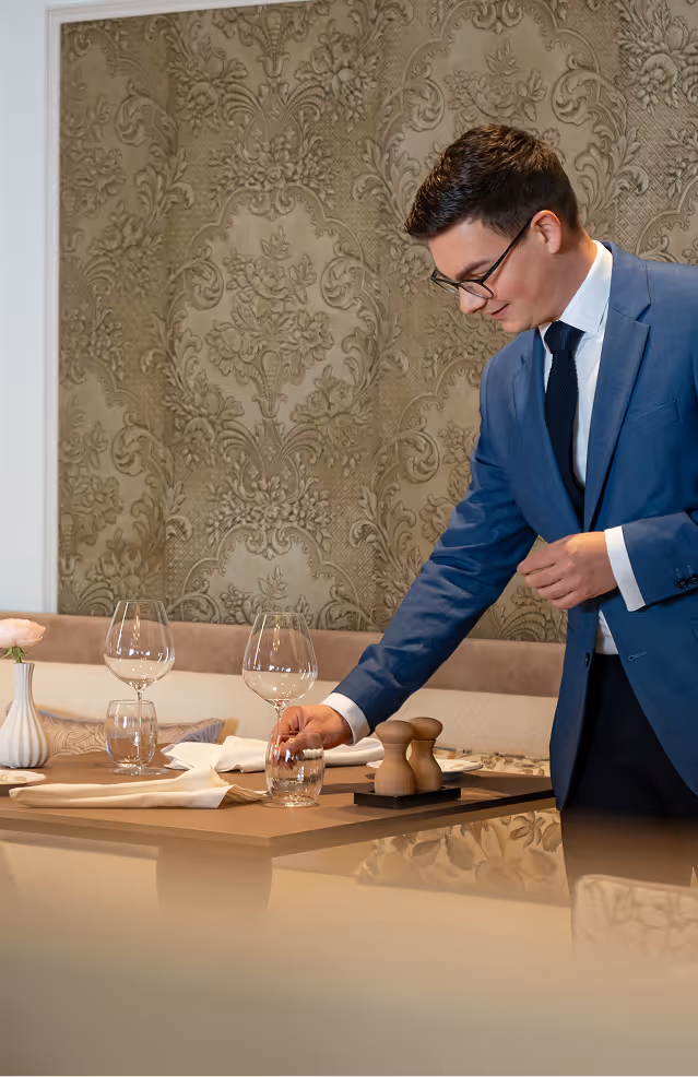Man in blue suit and glasses setting a glass on a restaurant table with wine glasses and napkins.
