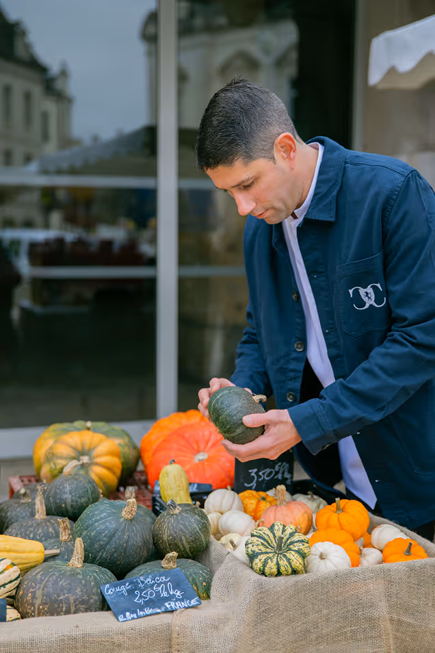 A man in a blue jacket inspecting a small green pumpkin at a market stall of various pumpkins.
