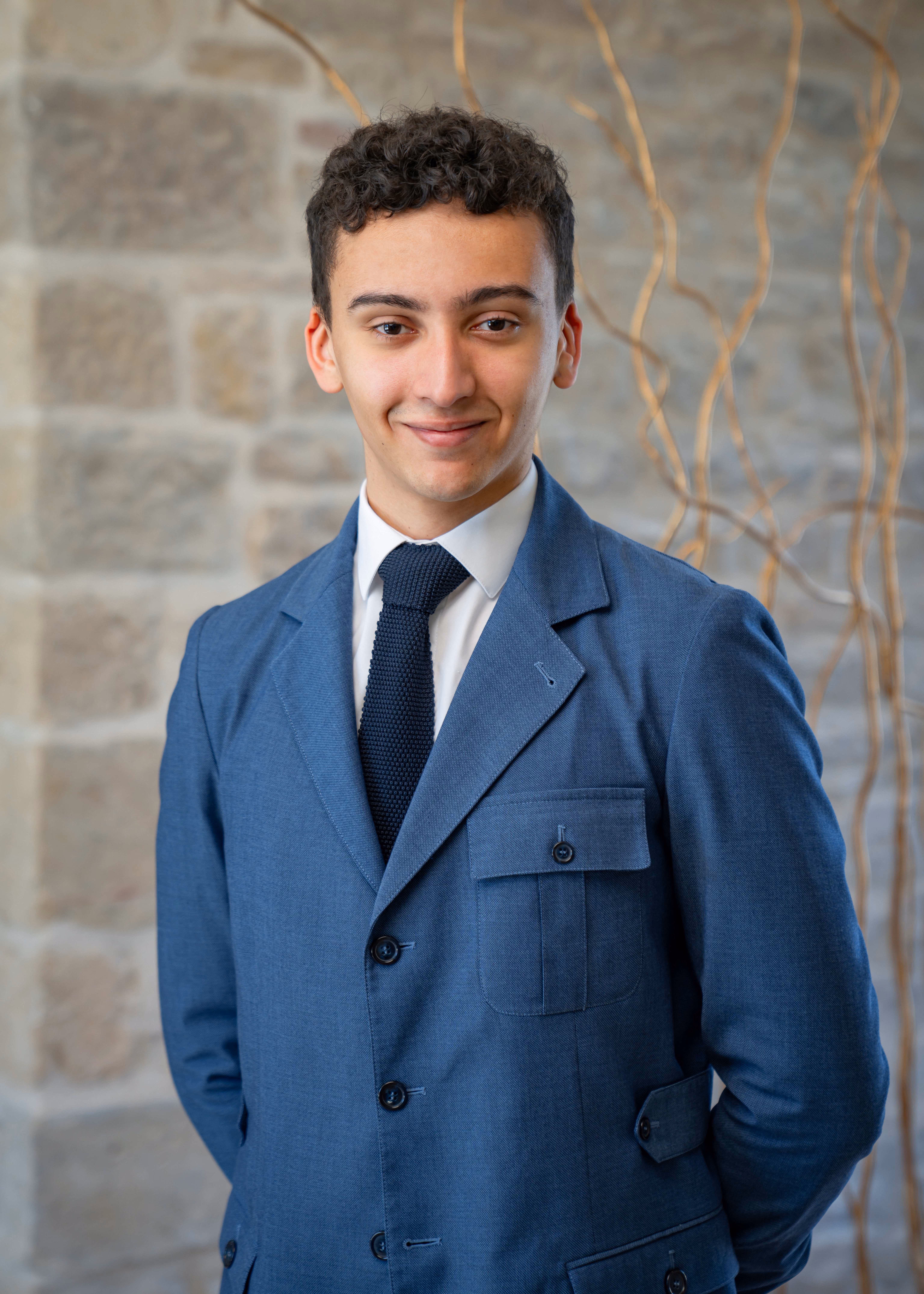 Young man with curly hair wearing blue suit and tie standing in front of a stone wall.
