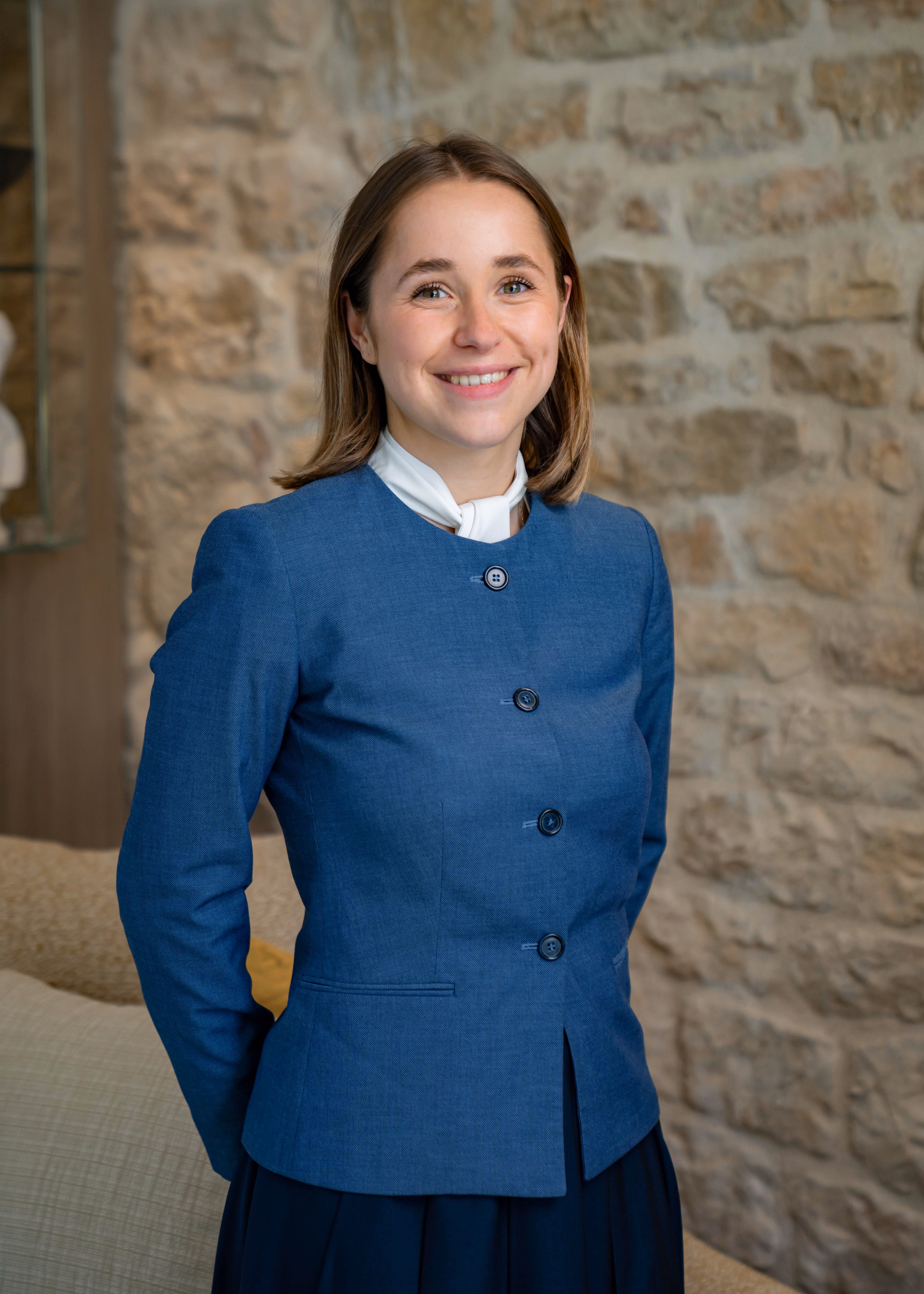 Smiling young woman with shoulder-length hair wearing a blue blazer and white scarf in front of stone wall.