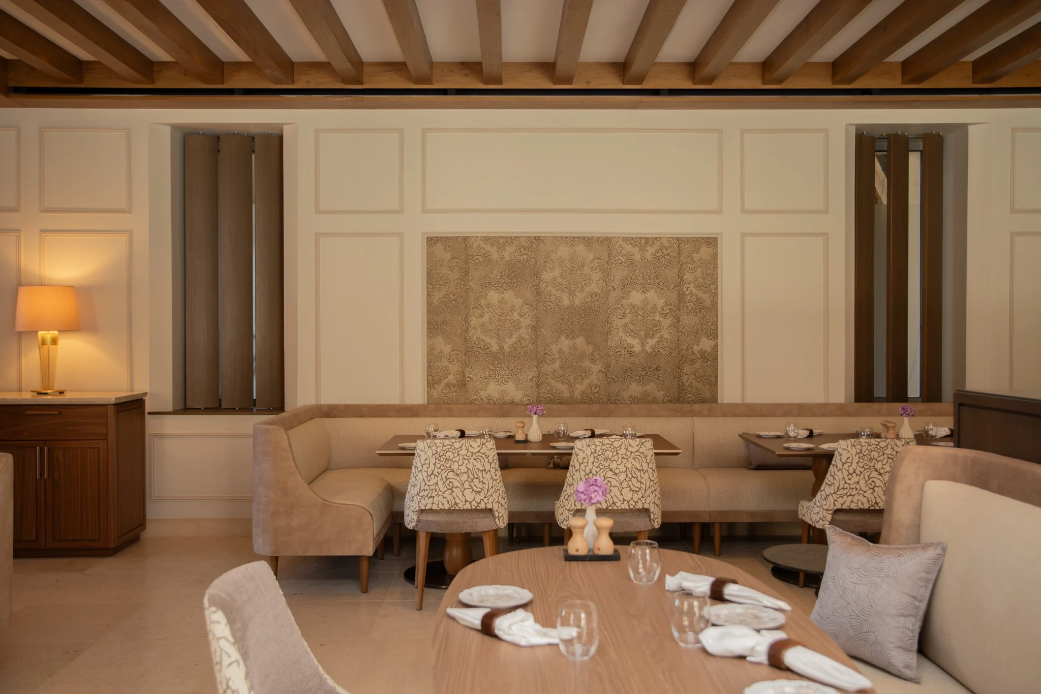 Elegant dining room with wooden ceiling beams, beige booths, and tables set with glassware and plates.