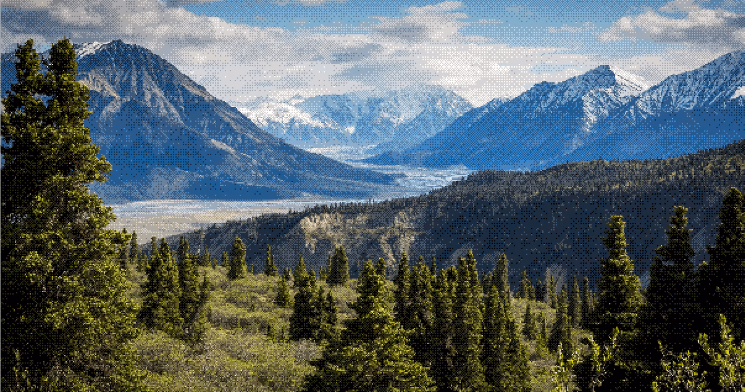 Green pine trees in the foreground with a mountain range and snow-capped peaks under a partly cloudy sky.